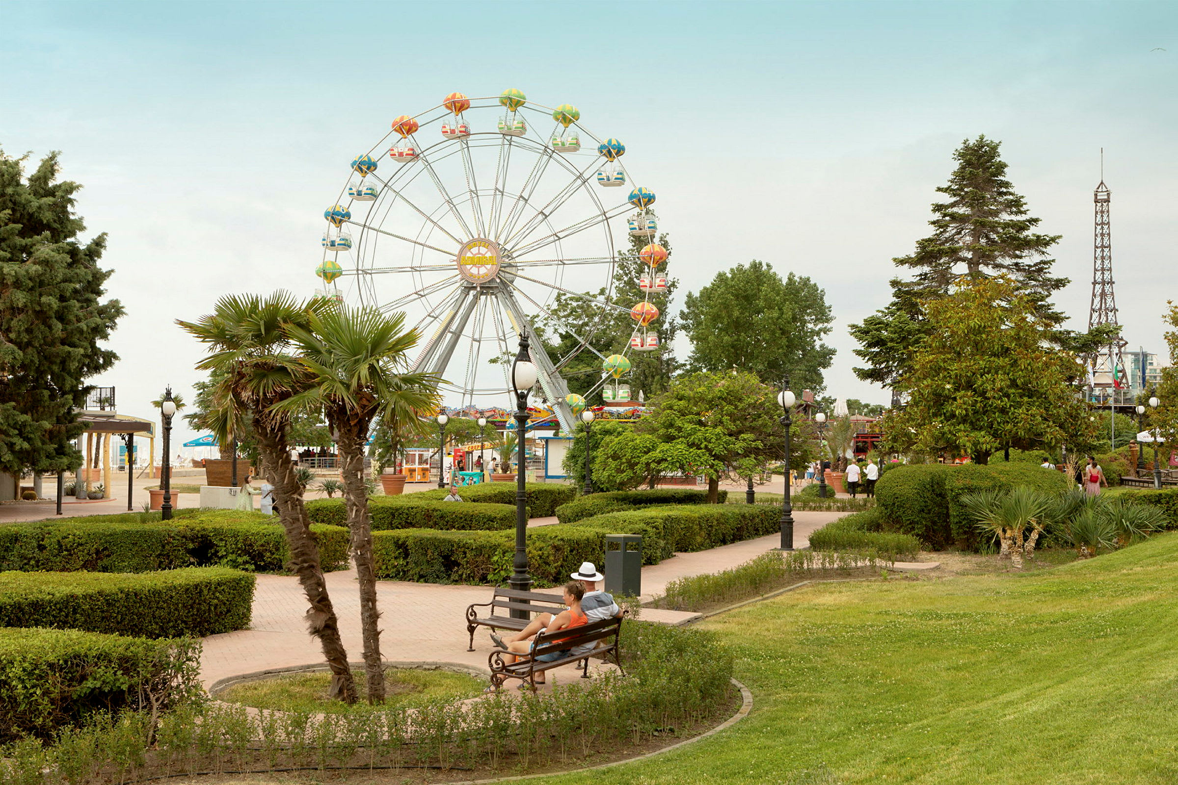 En park med gröna träd och buskar, en sittande person på en parkbänk, ett pariserhjul och en liten modell av Eiffeltornet i bakgrunden. (AI-genererad text)
