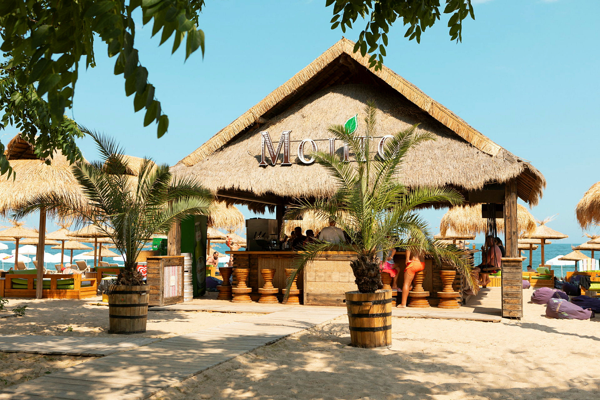 Strandbar med palmkvisttak omgiven av sand och solparasoller. (AI-genererad text)