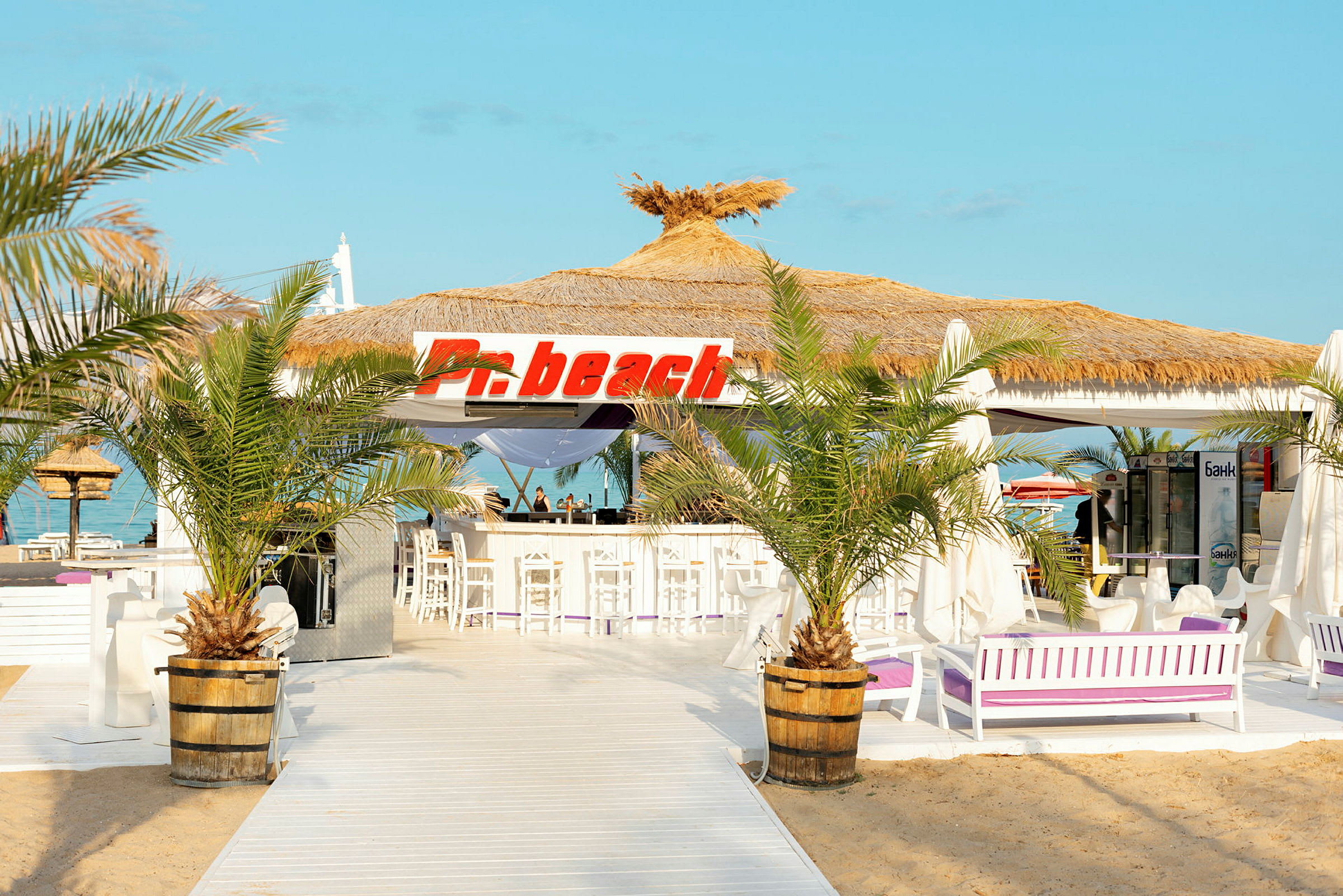 En strandbar med halmtak och skylt som säger 'Pn beach', omgiven av palmer och vit inredning. (AI-genererad text)