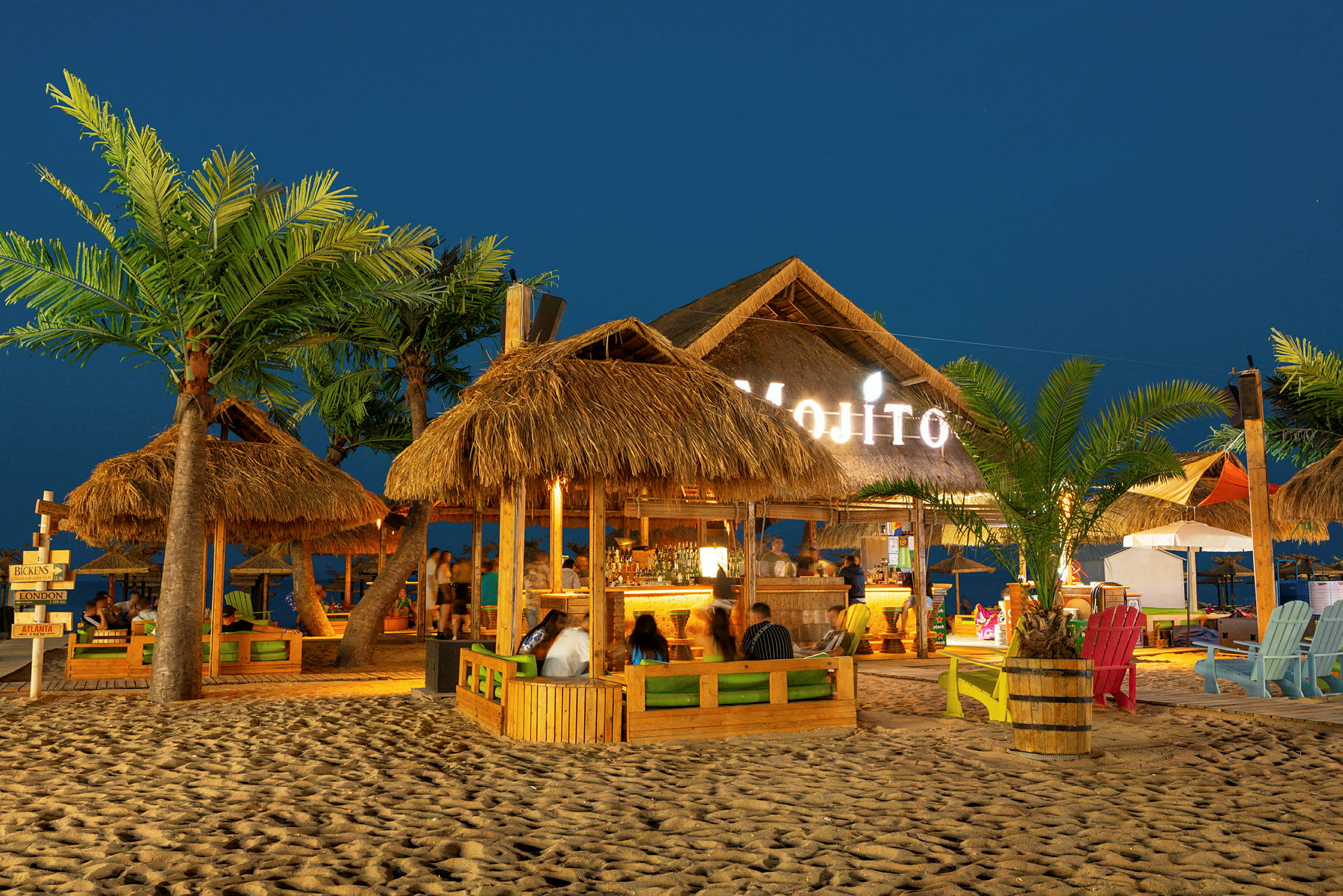 Strandbar med palmbladstak, stolar och bord på sandig mark på kvällen. (AI-genererad text)