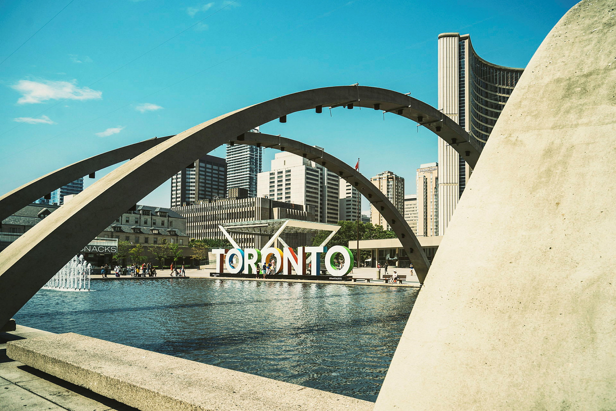 En vy över Nathan Phillips Square i Toronto med en fontän, bågar och stadens skyline i bakgrunden. (AI-genererad text)