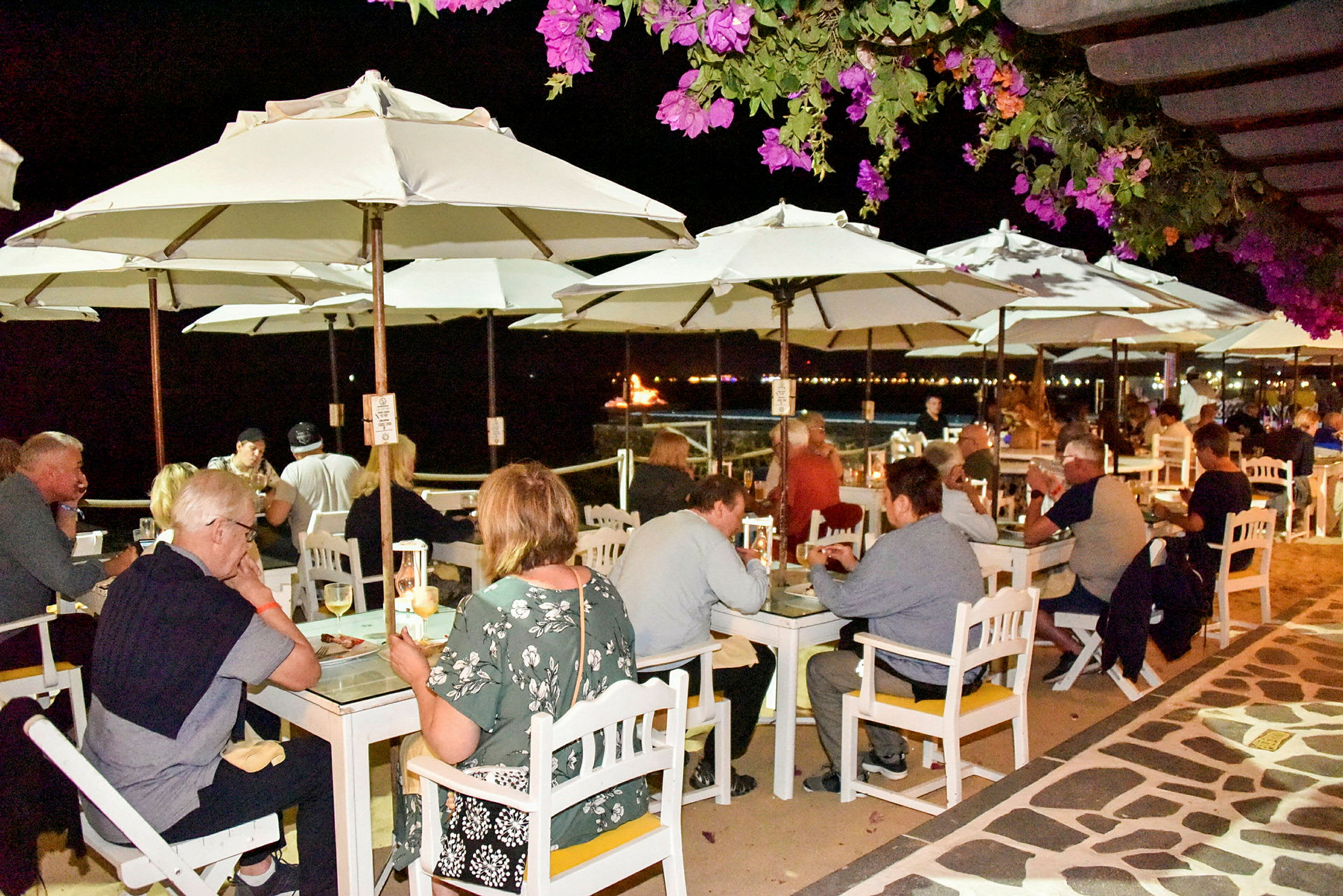Människor sitter vid utomhusbord under vita parasoller på en kvällsrestaurang, med blommande växter i förgrunden och havet i bakgrunden. (AI-genererad text)