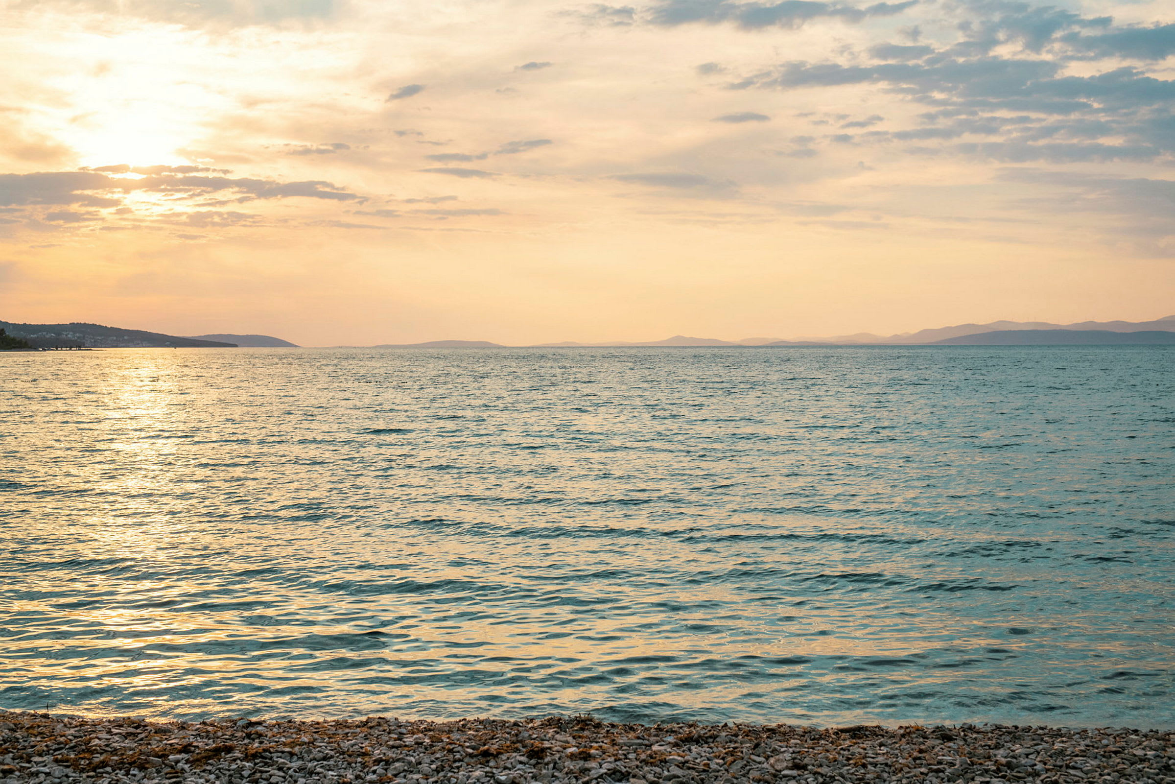 Strand med småsten vid havet i solnedgången med berg i bakgrunden. (AI-genererad text)