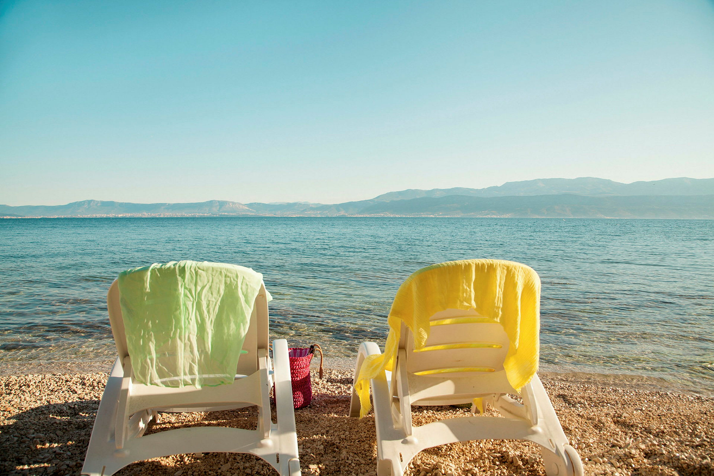 Två solstolar med handdukar på en stenstrand med utsikt över havet och berg i bakgrunden. (AI-genererad text)
