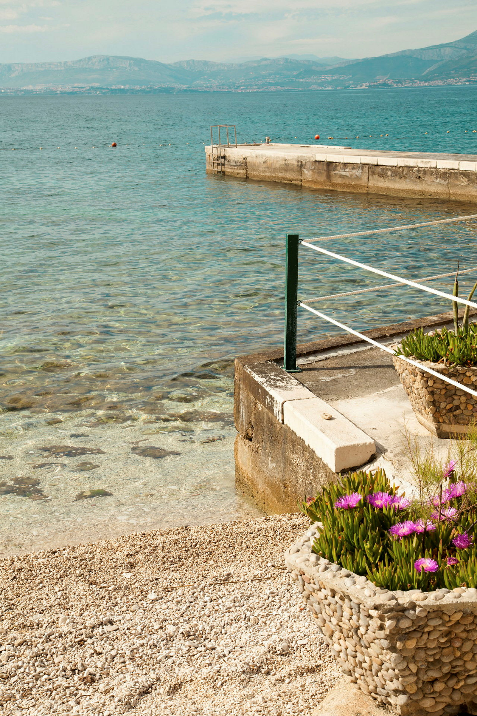 Strand med sten, blomkruka med lila blommor och brygga vid klart vatten. (AI-genererad text)