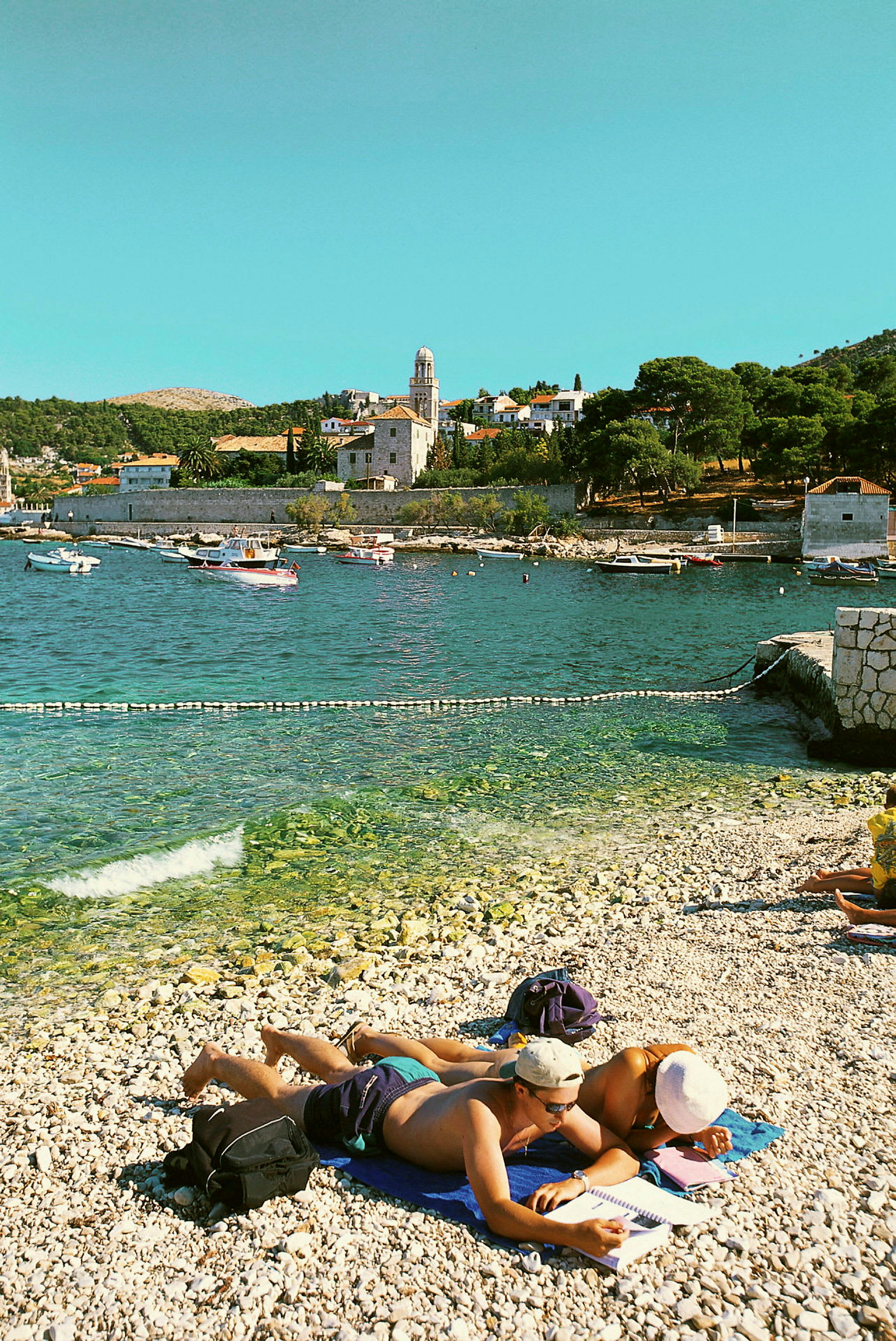 Personer som ligger på en stenstrand nära havet med soliga byggnader i bakgrunden. (AI-genererad text)