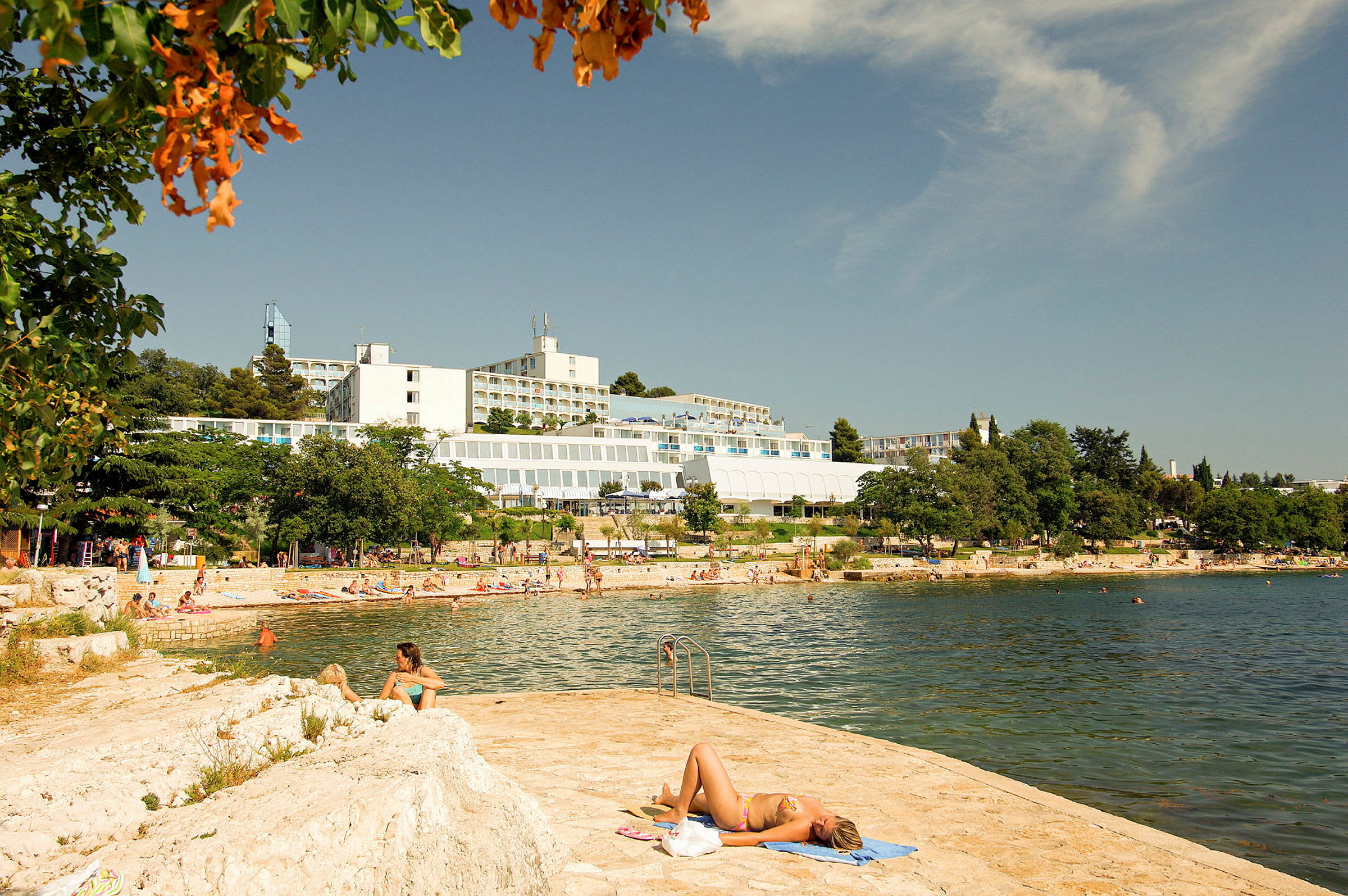 En person ligger på en handduk vid vattnet med hotellbyggnader och en strand i bakgrunden. (AI-genererad text)