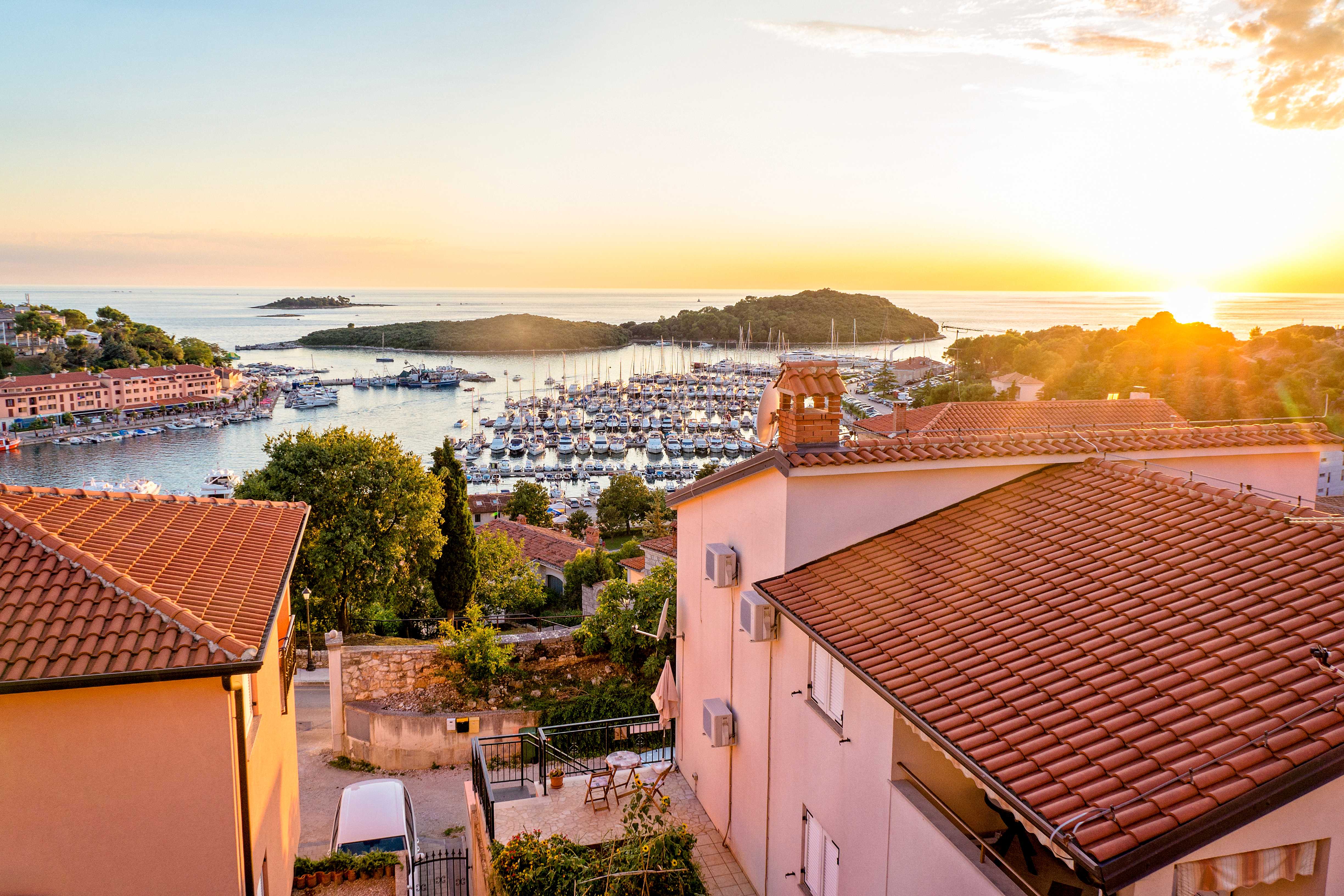Vy över en småbåtshamn med segelbåtar omgivna av hus och grönska vid solnedgång. (AI-genererad text)