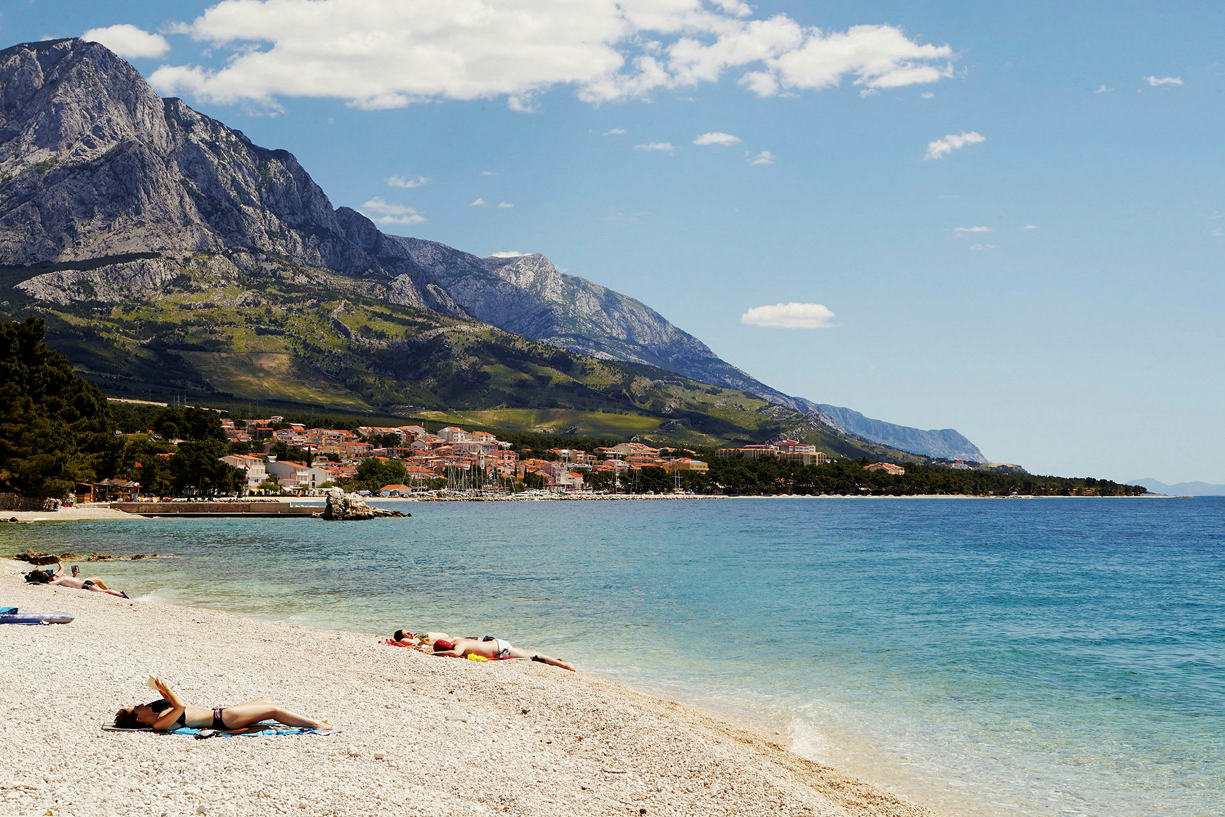 En strandscen med personer som solar, kuststad i bakgrunden och berg. (AI-genererad text)