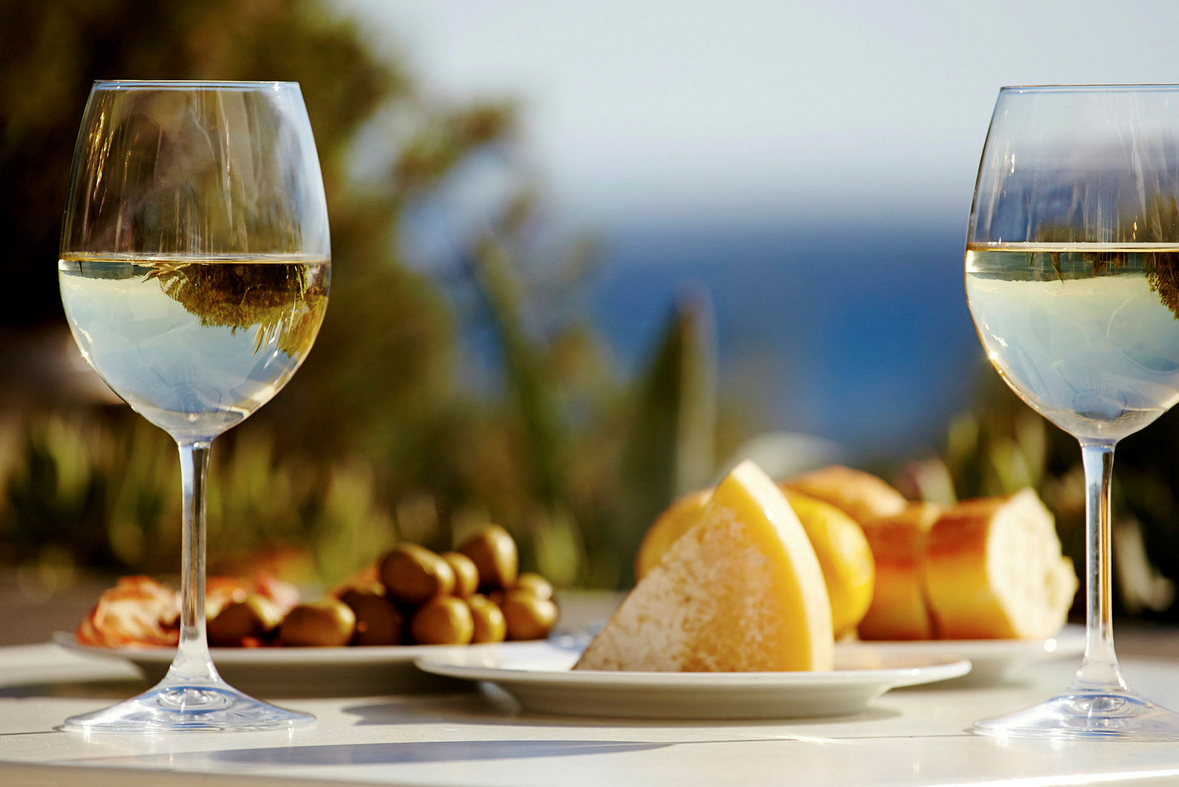 Två glas vitt vin med en tallrik innehållande ost, bröd och oliver i bakgrunden. (AI-genererad text)