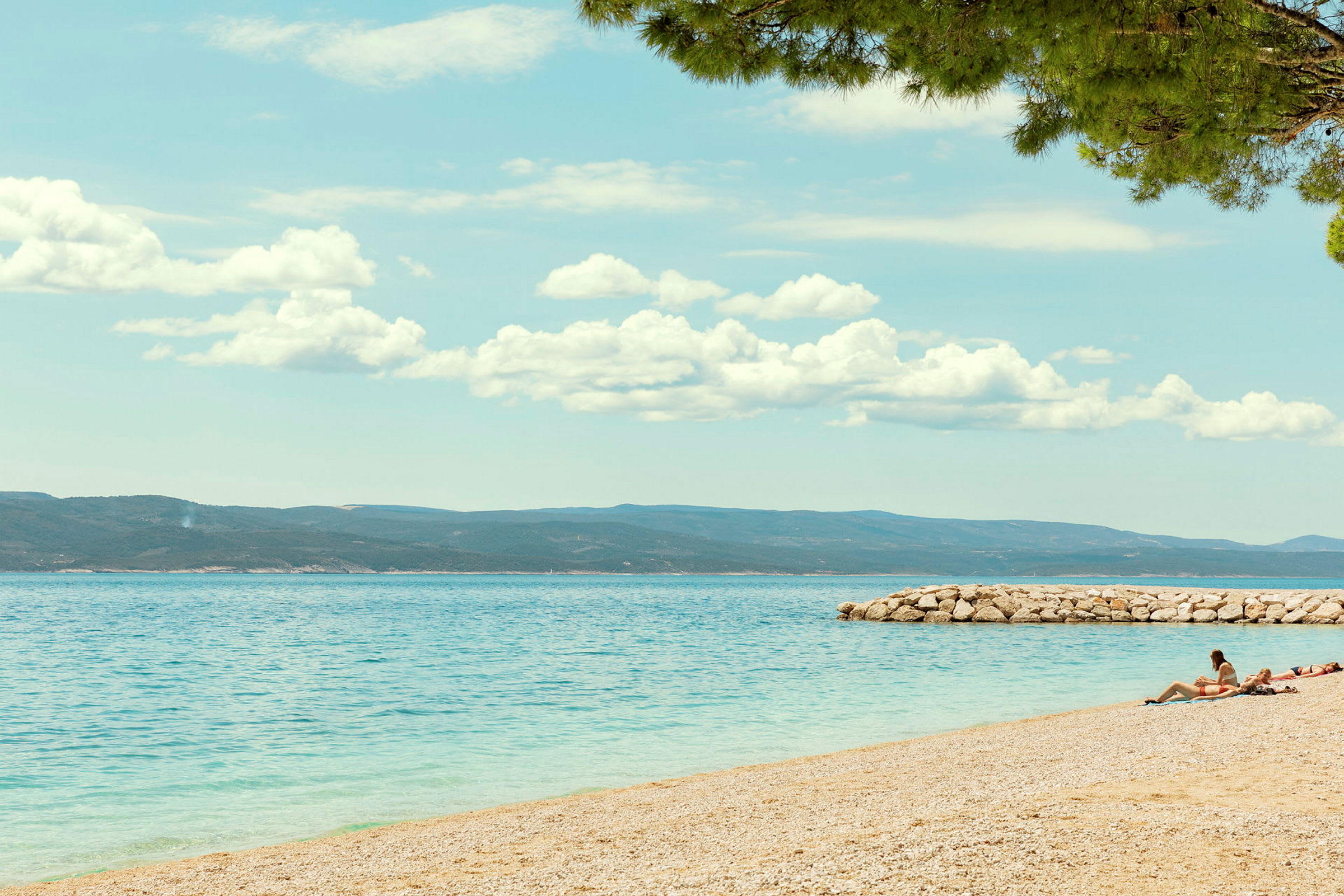Strand med klarblått vatten och personer som solar vid en stenpir. (AI-genererad text)