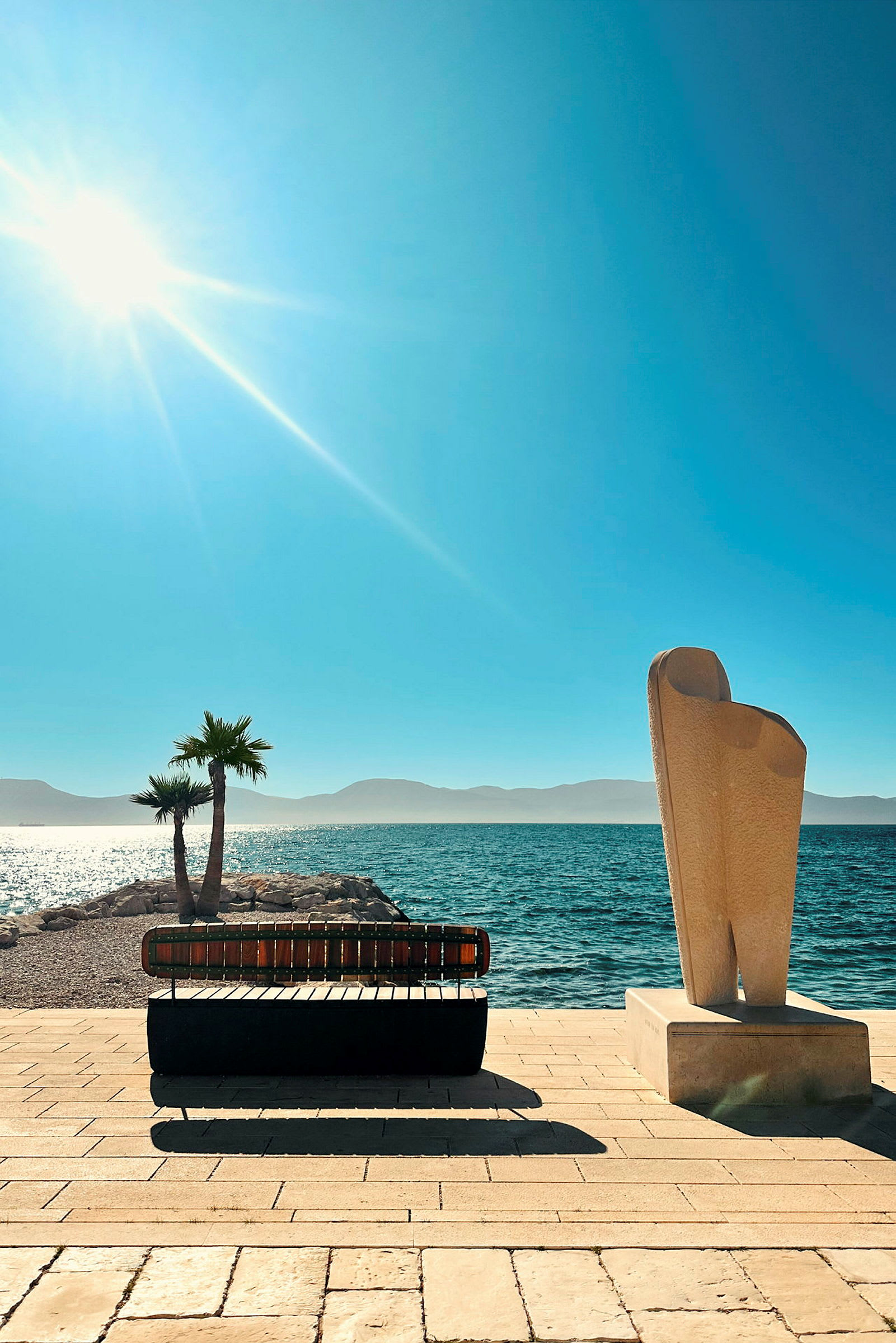 En strandpromenad med en solbänk och en skulptur, med havet och solen i bakgrunden. (AI-genererad text)