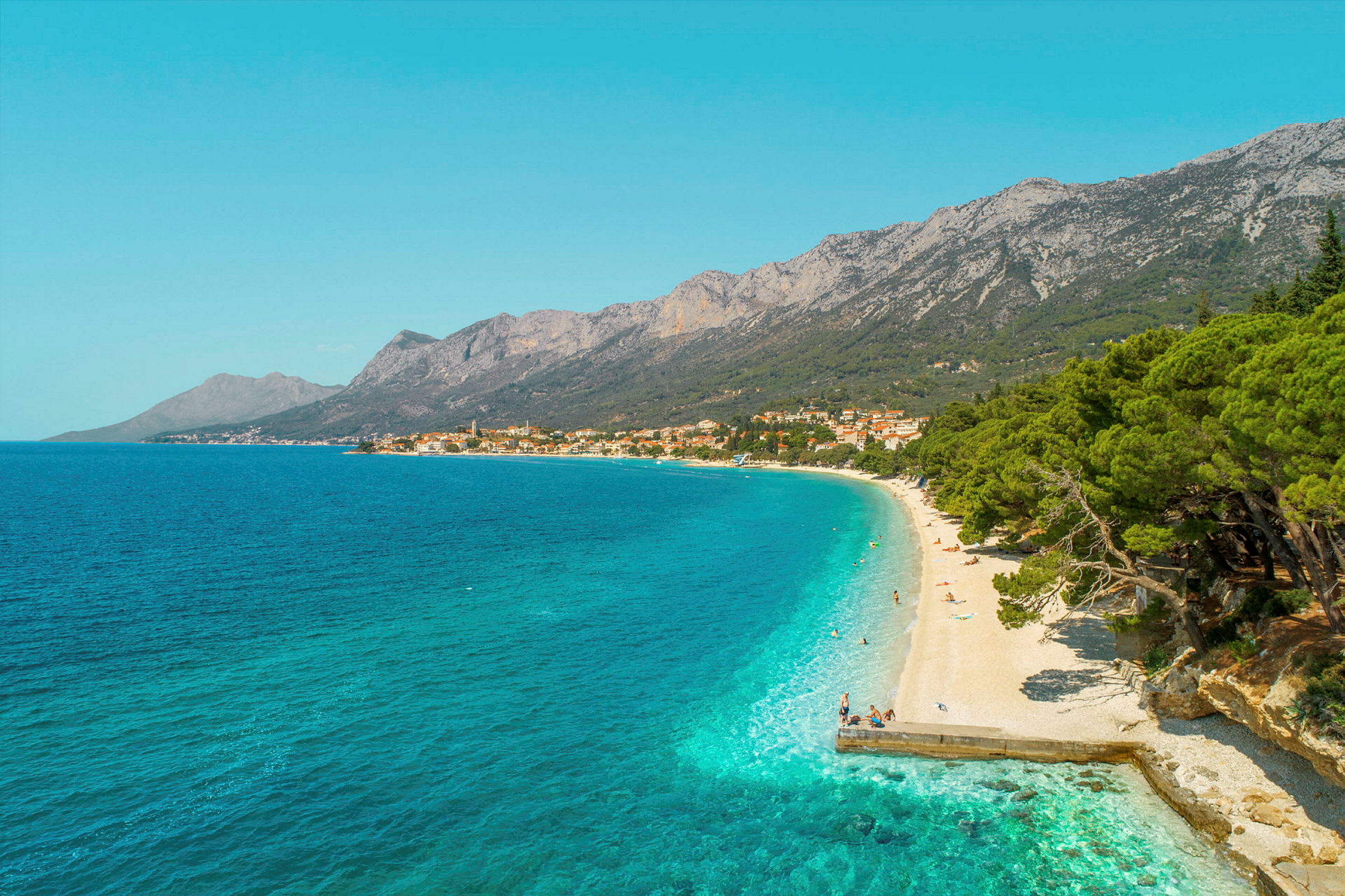 Vy över en klapperstensstrand med turkost vatten och grönskande träd. Berg och en by syns i bakgrunden. (AI-genererad text)