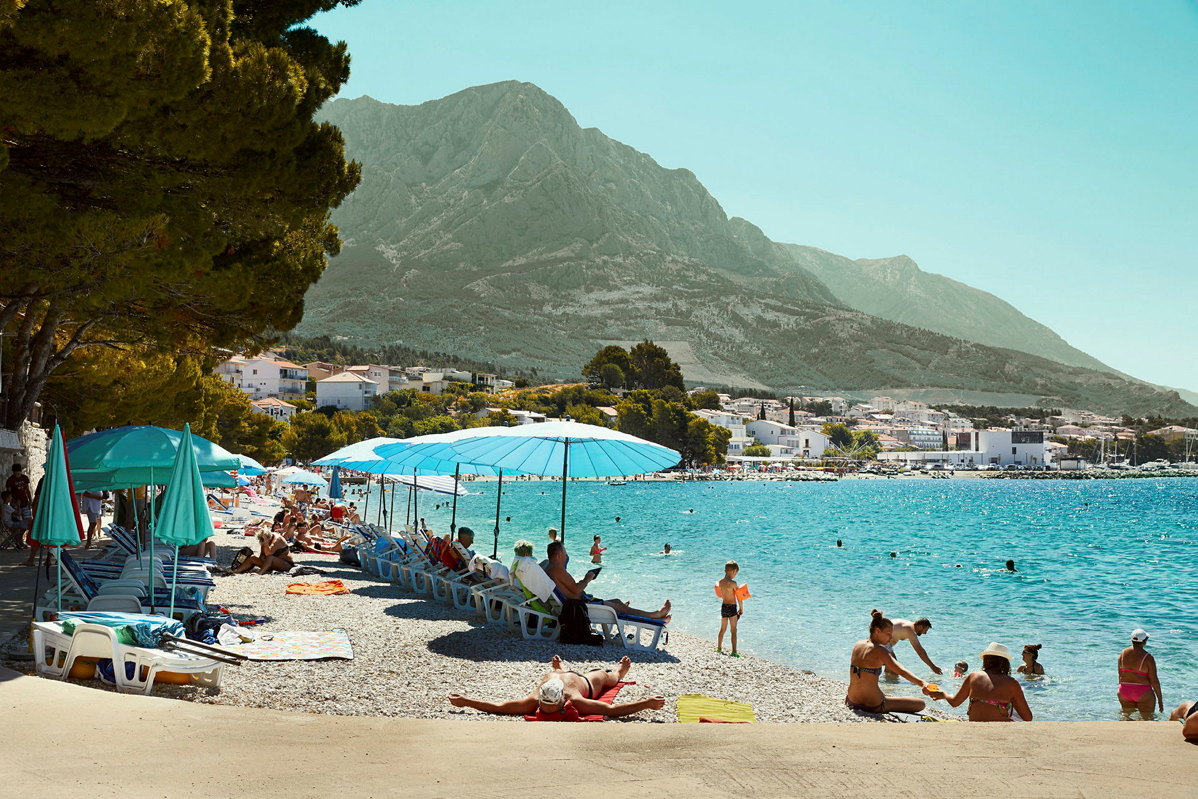 Strand med paraplyer och solsängar vid klart vatten, omgiven av berg. (AI-genererad text)