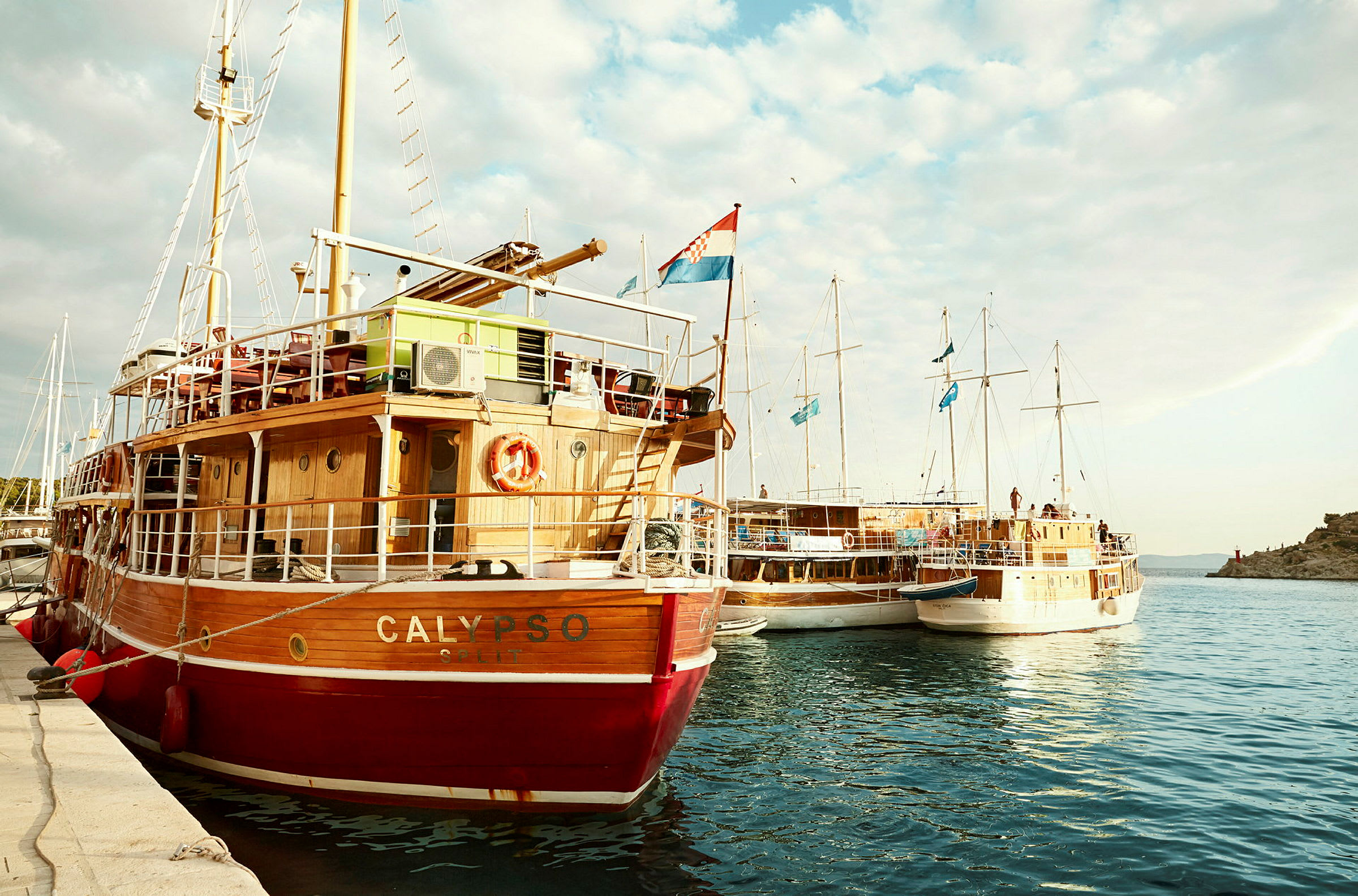 En röd och vit segelbåt med namnet Calypso vid en brygga, omgiven av andra båtar med blå flaggor. (AI-genererad text)