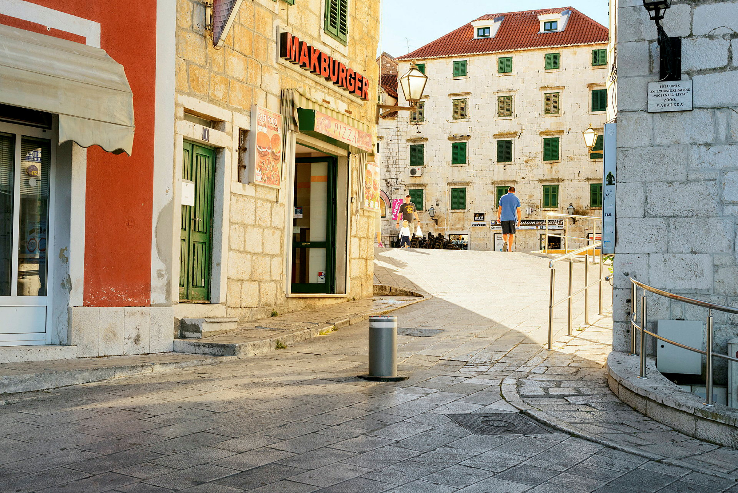 Stadsmiljö med stengata och byggnader med röda tak och gröna fönsterluckor samt en restaurangskylt med texten 'Makburger'. (AI-genererad text)