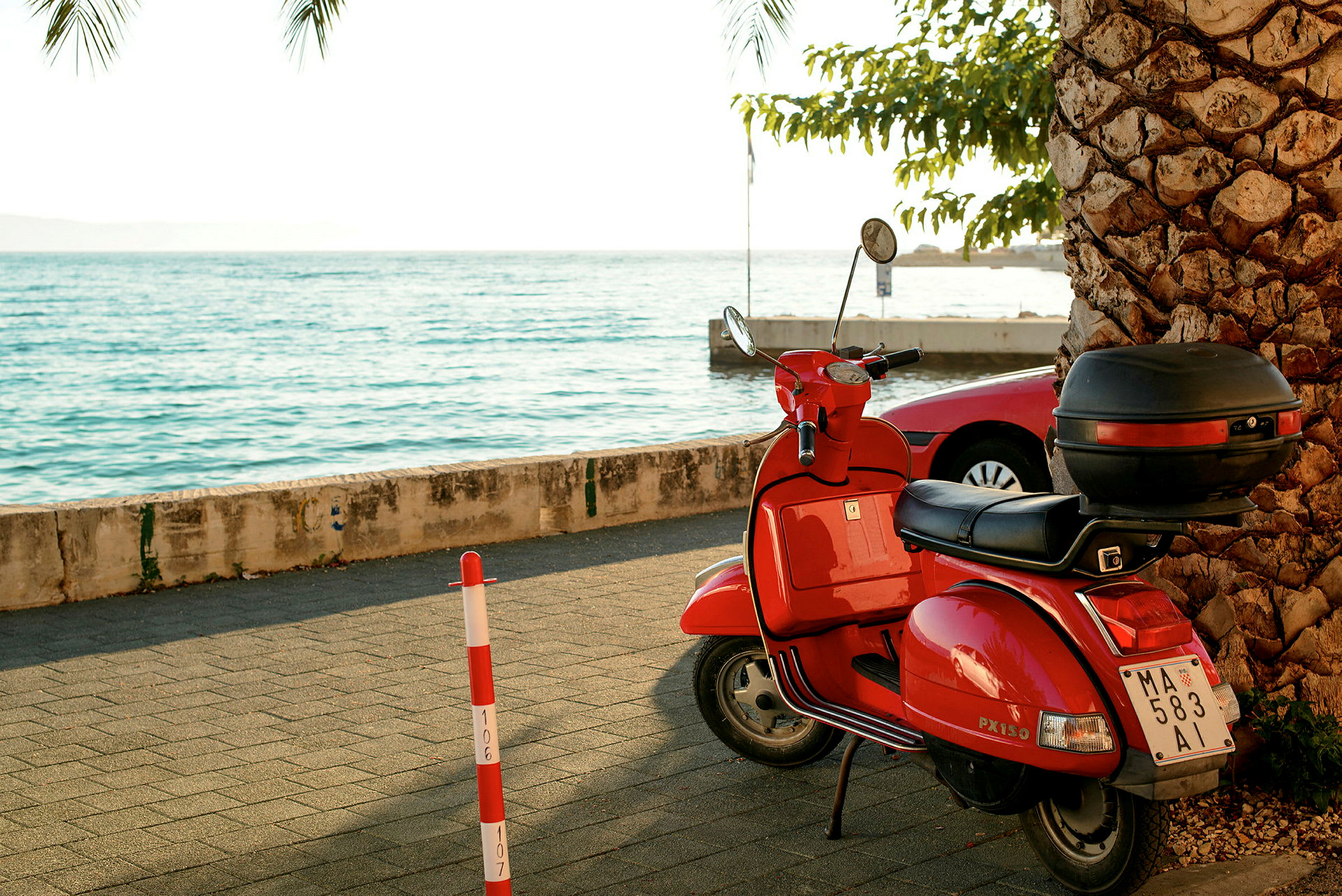 Röd moped parkerad på gångväg nära havet, omgiven av en palm och parkerade bilar. (AI-genererad text)