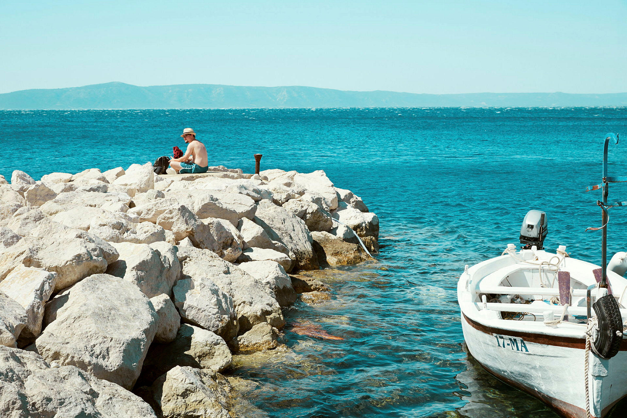En person sitter på klippor nära havet, med en liten båt förtöjd vid sidan. (AI-genererad text)