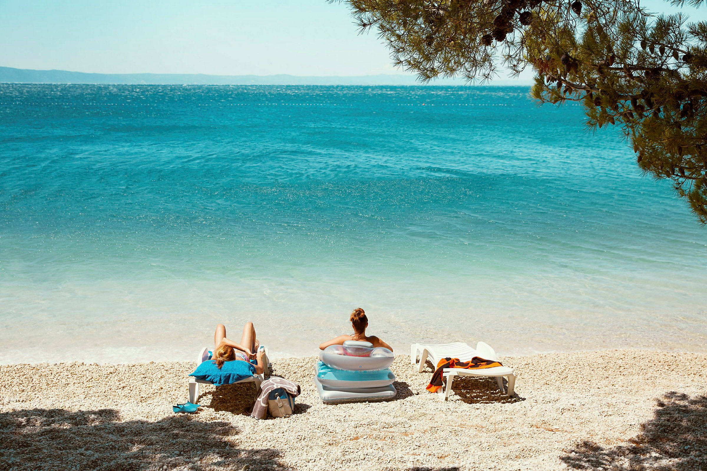 Två personer sitter i solstolar på en strand med blått hav och omgivande grönska. (AI-genererad text)