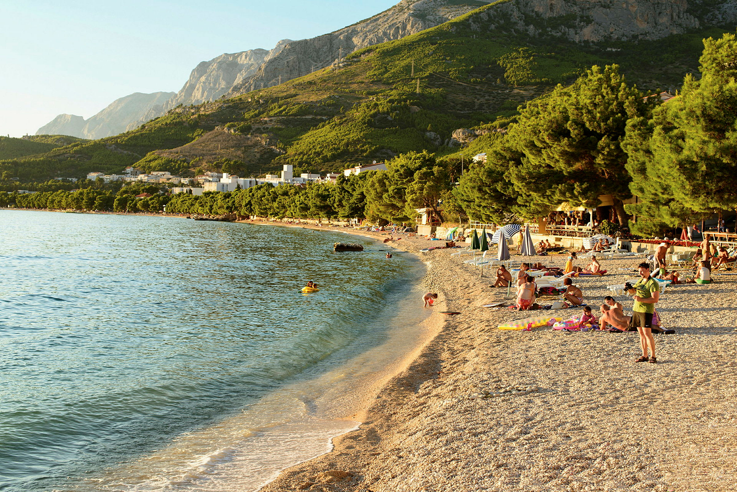 Strand med klappersten och badande människor omgiven av grönska och berg i bakgrunden. (AI-genererad text)