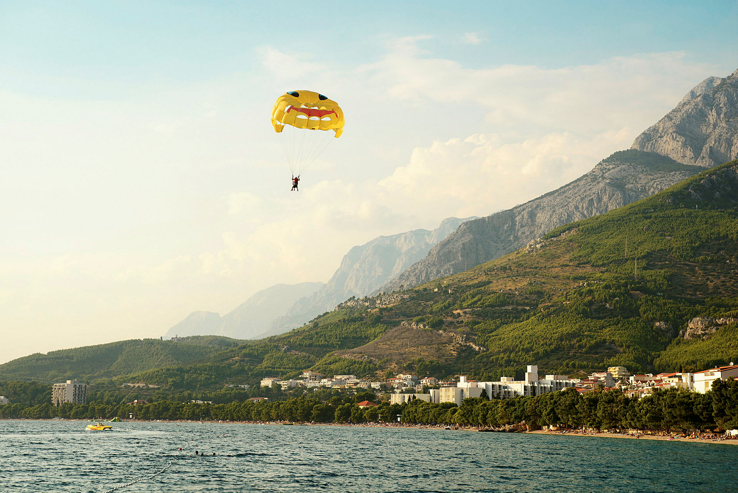 En person paraglider med ett fallskärm med ansiktsmotiv över en kustlinje med berg i bakgrunden. (AI-genererad text)