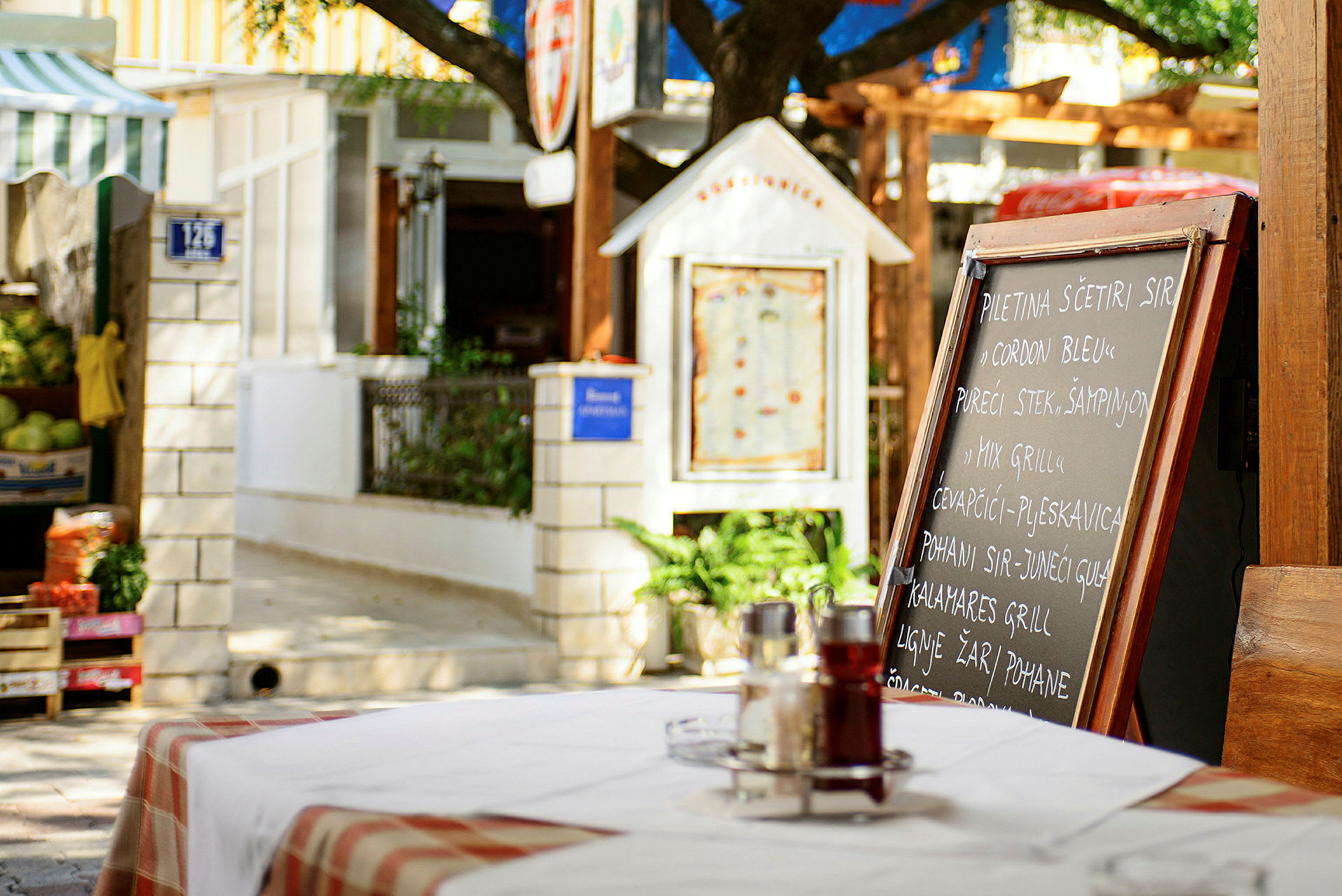 En restaurangmeny på en svart tavla utomhus på ett bord med en rödvit duk, omgiven av grönska och byggnader. (AI-genererad text)