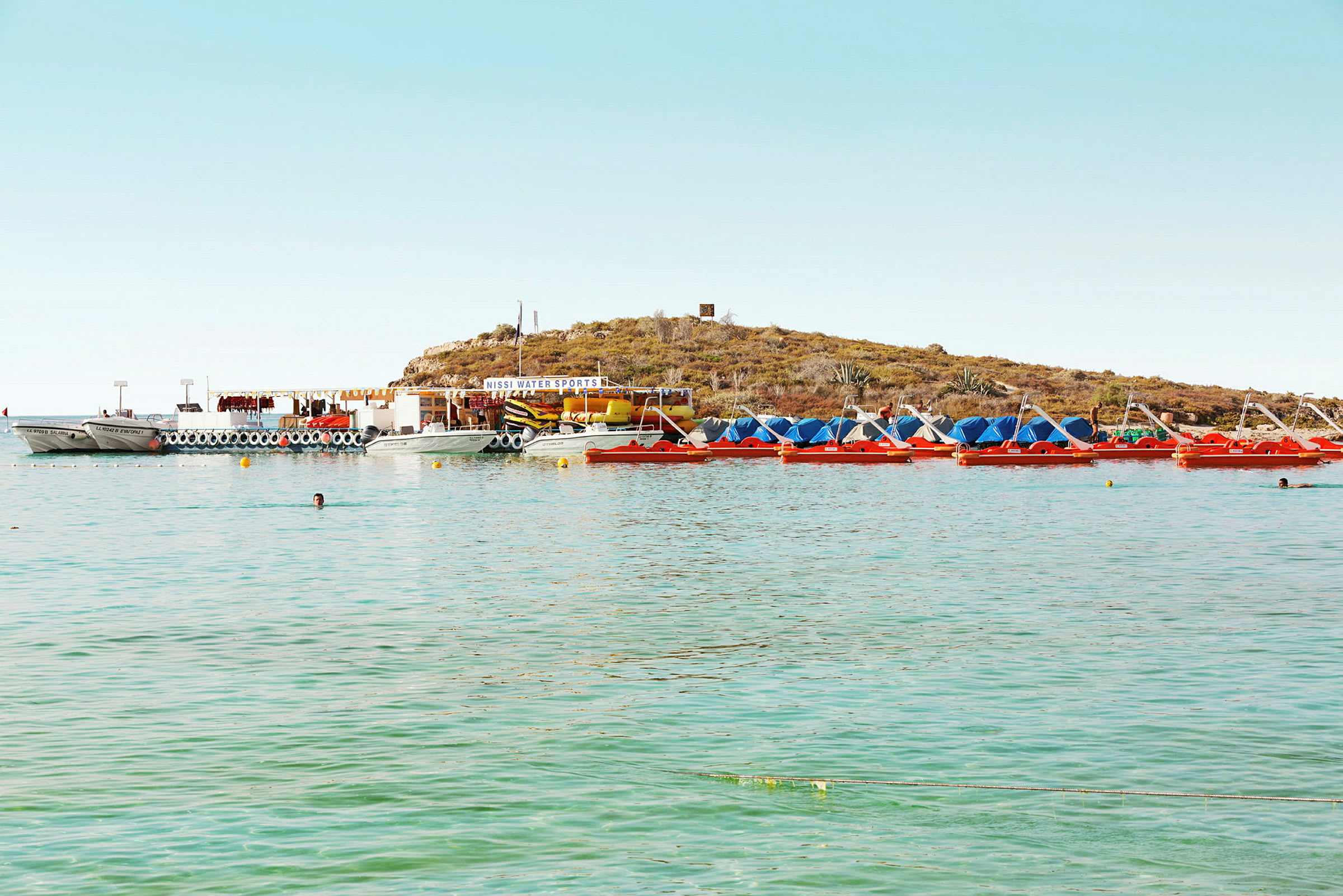 En samling av båtar och trampbåtar vid en strand med gröna vatten och en liten kulle i bakgrunden. (AI-genererad text)
