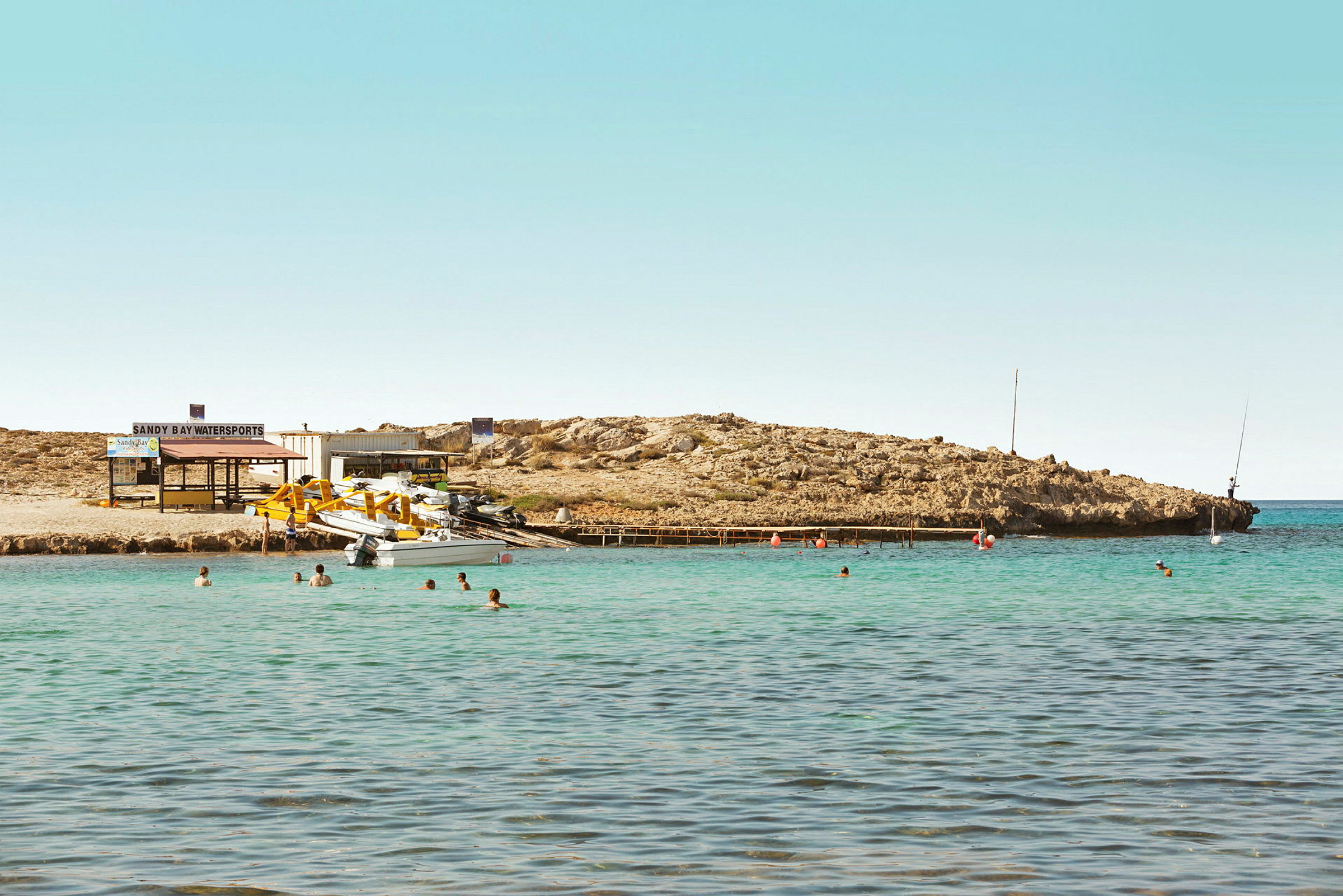 En liten vik med klart vatten. Människor badar och båtar är dockade vid en brygga. En vattensportanläggning med utrustning är belägen vid stranden. (AI-genererad text)