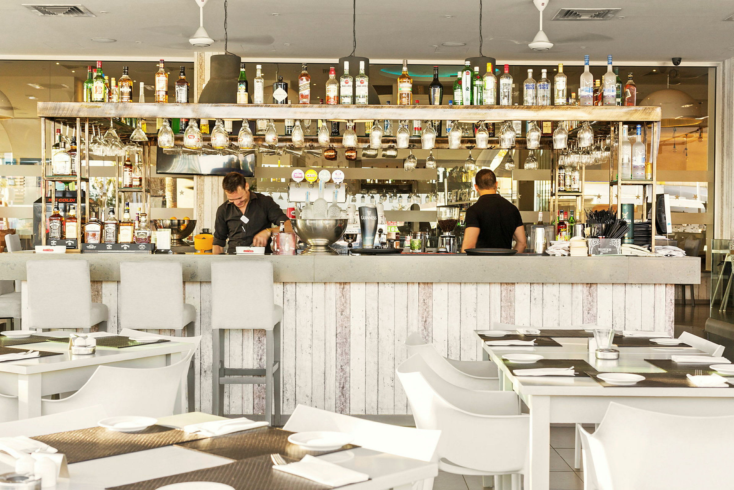 En välutrustad bar med cocktailglas hängande och dryckesflaskor på hyllan, med sittplatser och dukade bord framför. (AI-genererad text)
