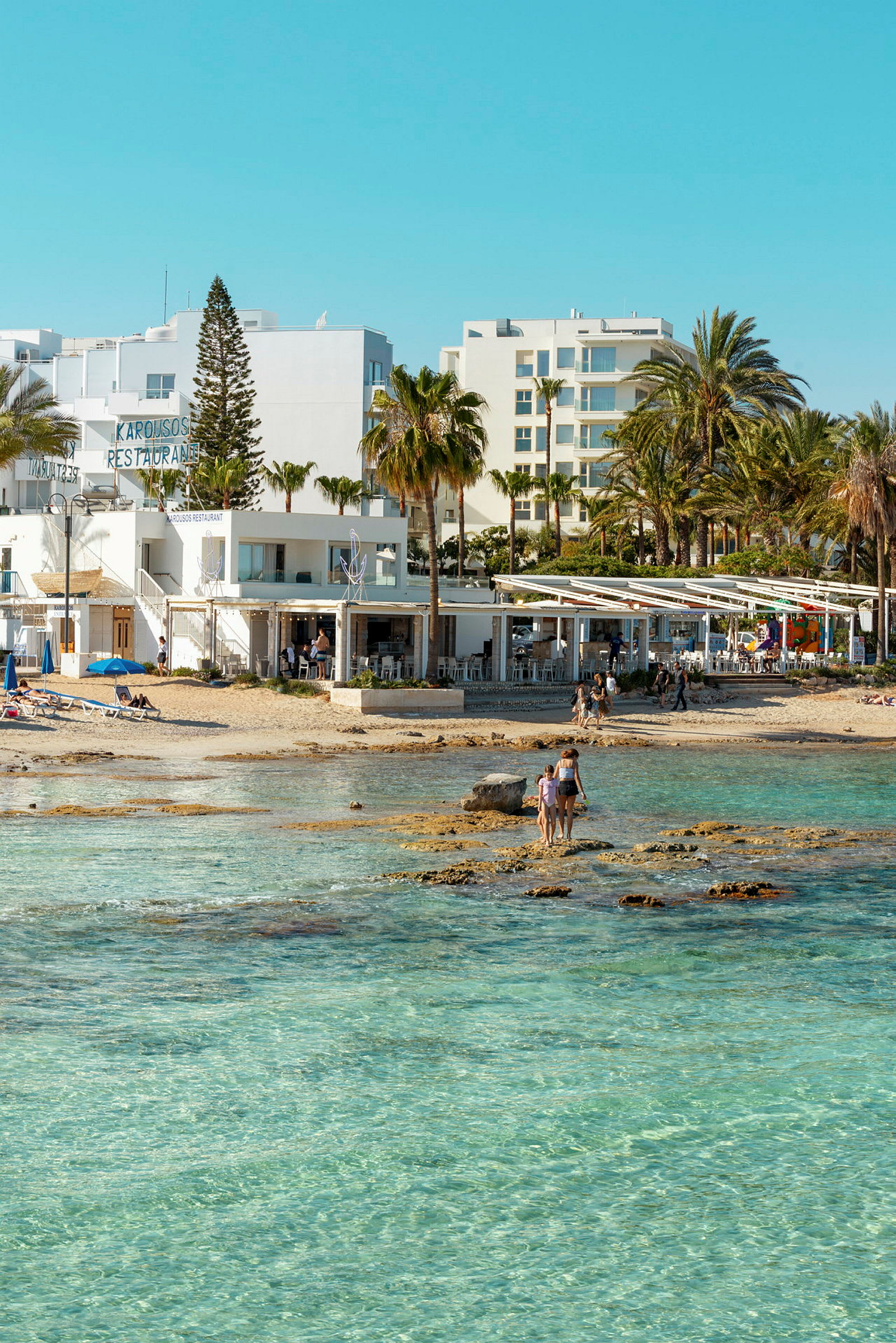 Strand med kristallklart vatten, byggnader och palmer i bakgrunden. (AI-genererad text)