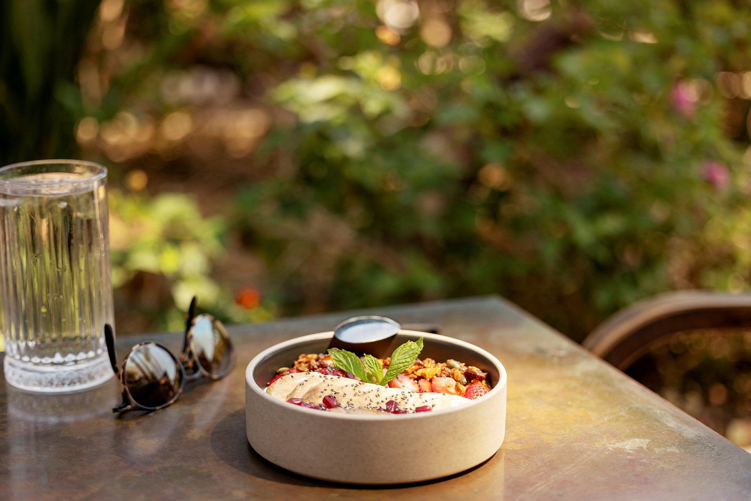 En skål med frukosttillbehör på ett bord tillsammans med ett glas vatten och solglasögon utomhus. (AI-genererad text)