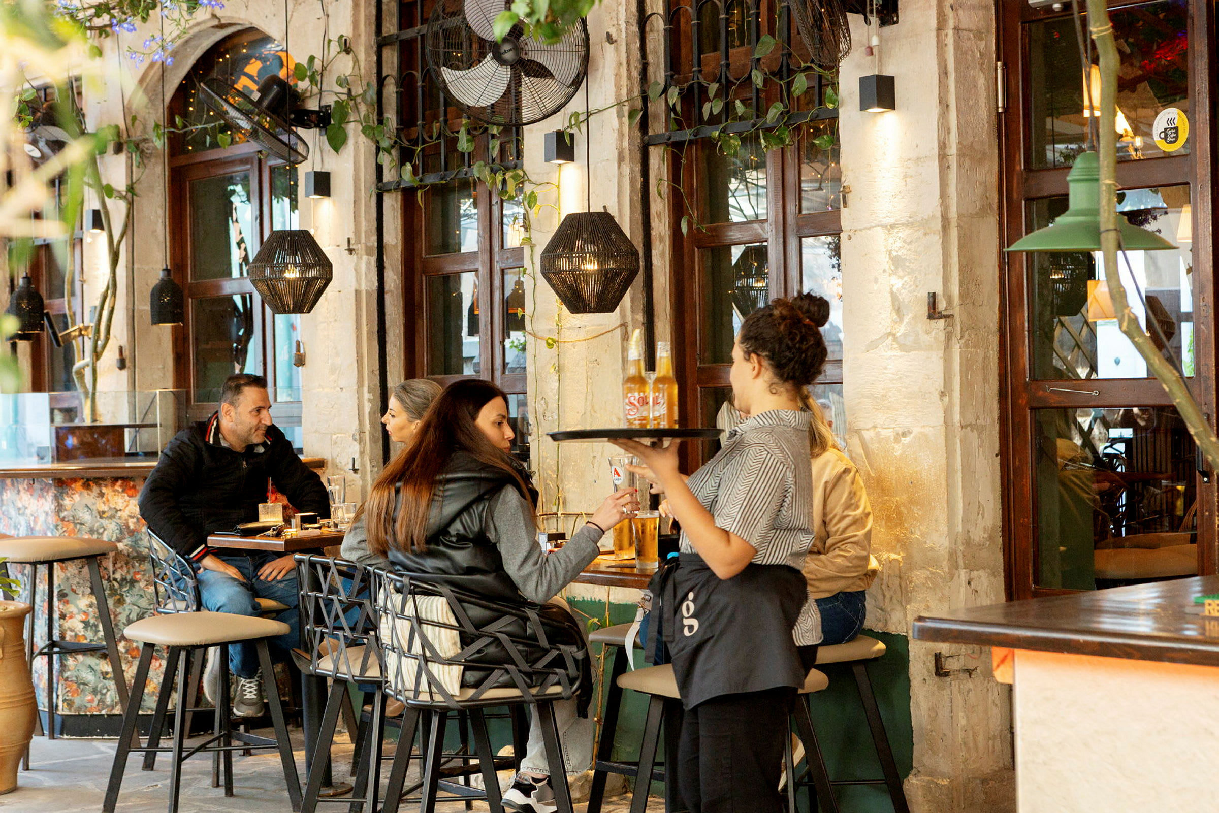En servitris serverar drycker till gäster i en restaurang med tegelväggar och fönster. (AI-genererad text)