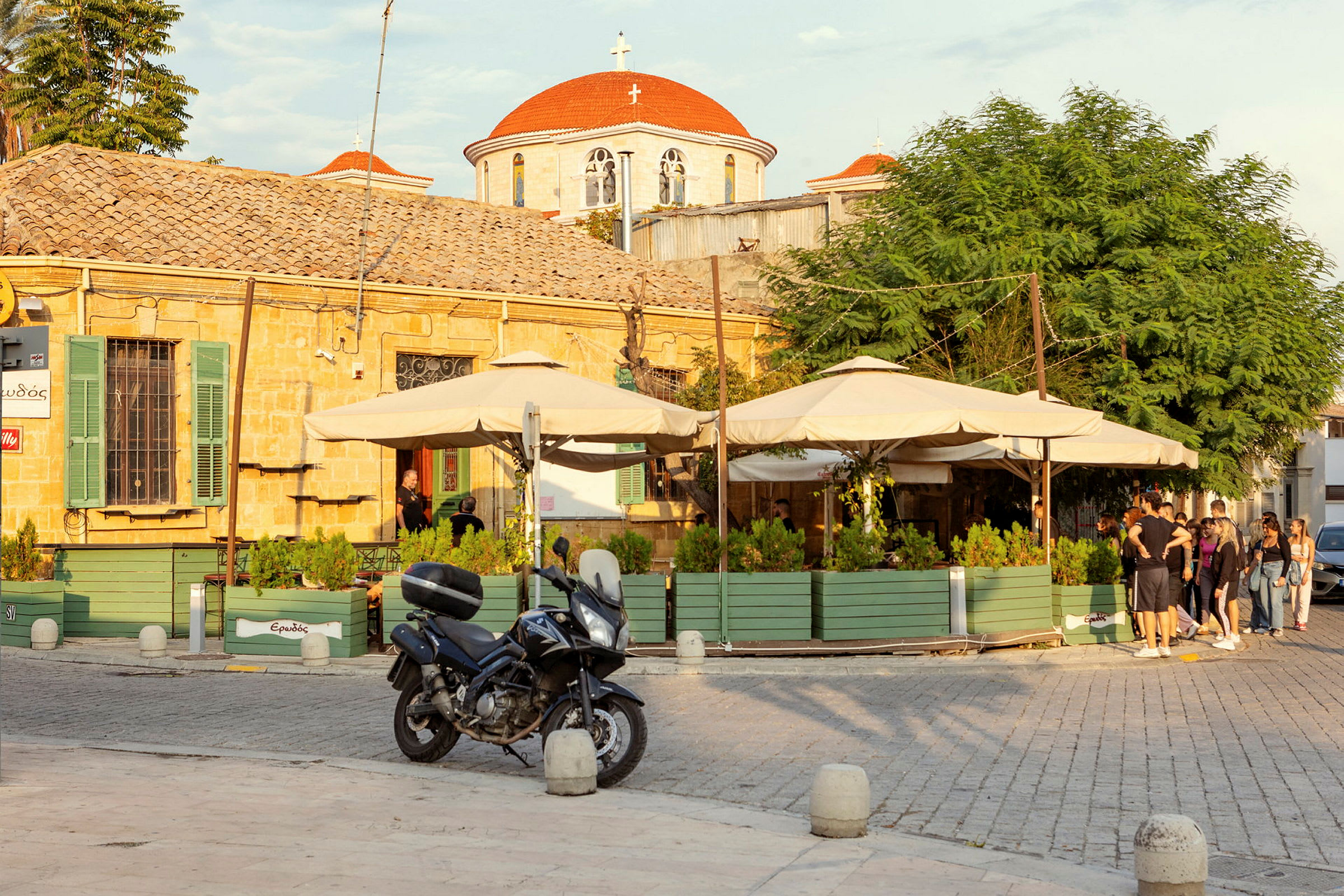 Ett café med gröna trälådor och parasoller framför en byggnad i sten med rött tak. En motorcykel står parkerad. (AI-genererad text)