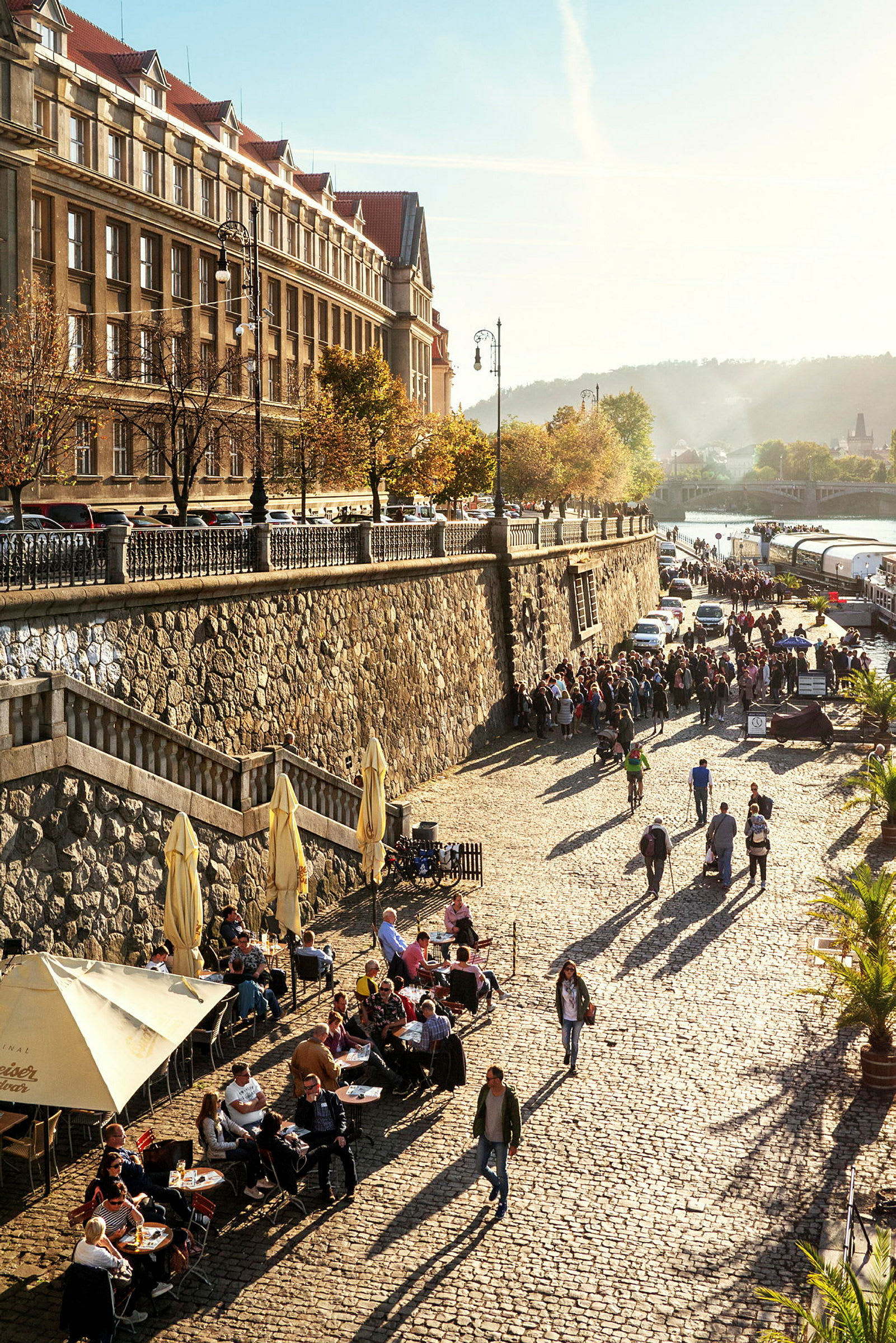 Gatubild med kullerstensgata vid en vattensamling, omgiven av en stenmur och byggnader, med människor som promenerar och sitter vid bord. (AI-genererad text)