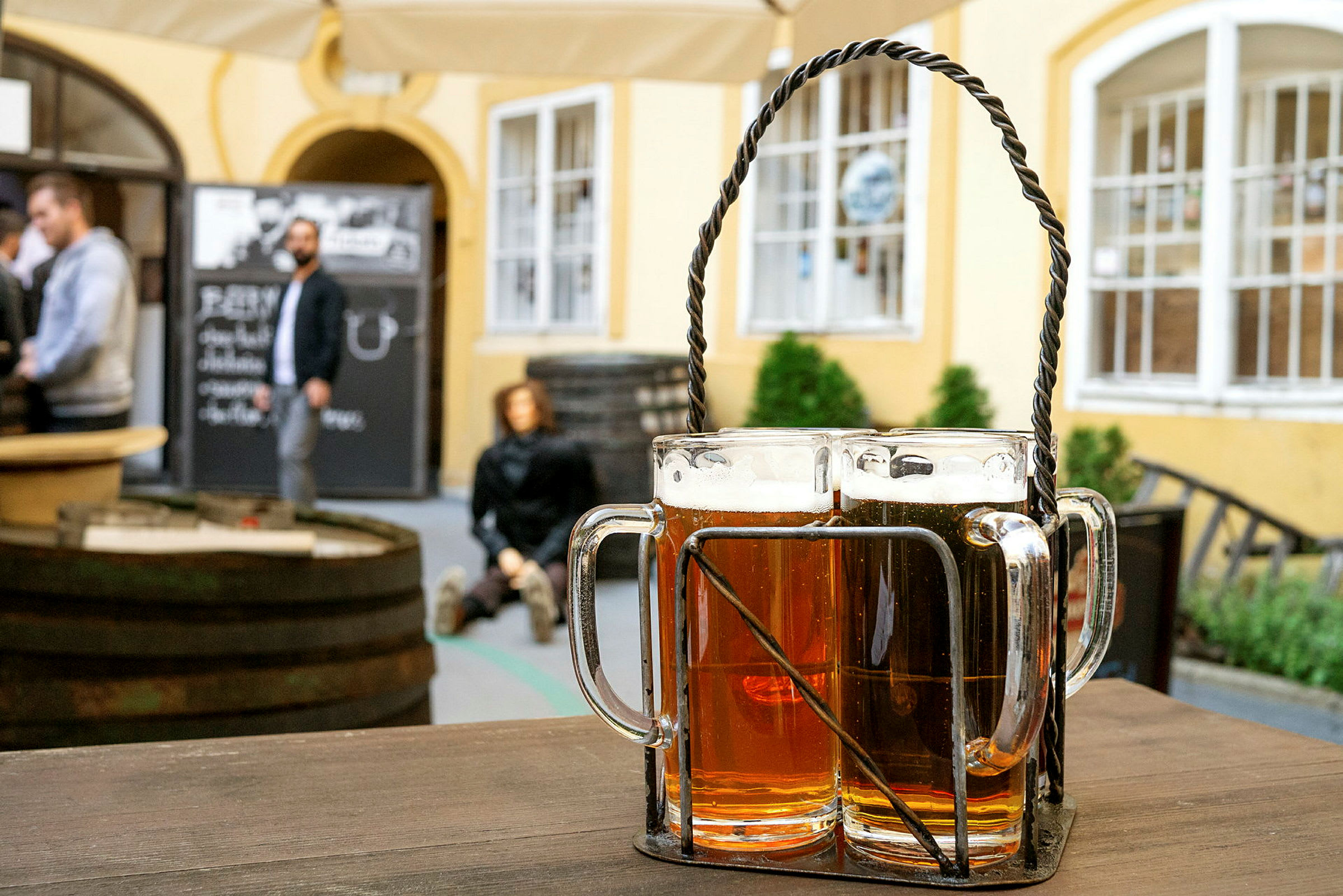 Två ölglas i hållare står på ett träbord i en utomhusrestaurang med personer i bakgrunden. (AI-genererad text)