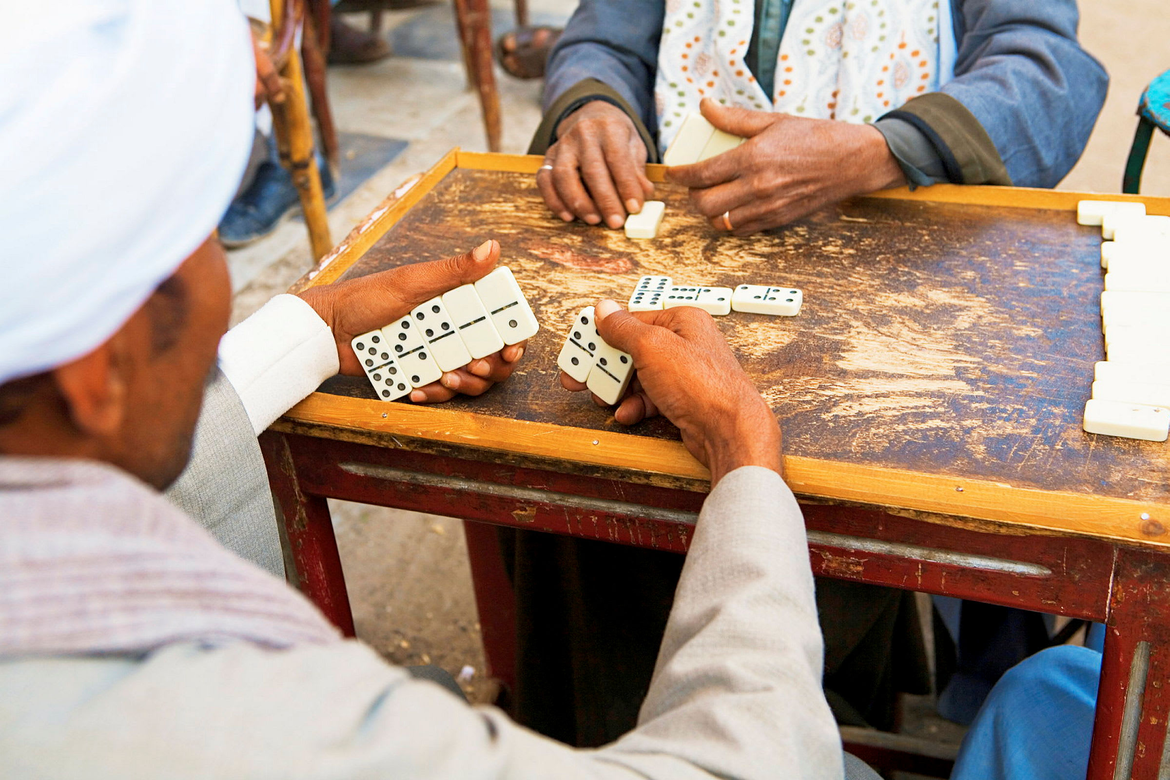 Två personer spelar domino vid ett bord, där den ena håller brickor i handen. (AI-genererad text)