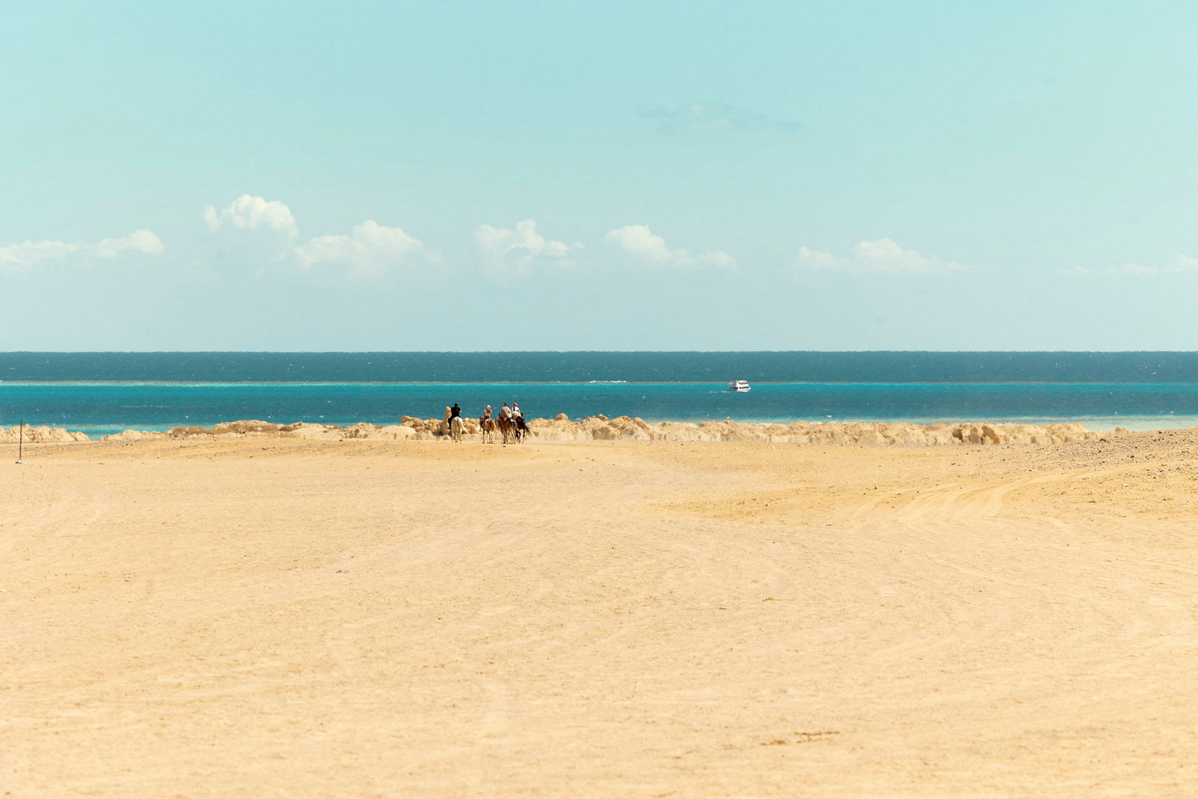 Ökenlandskap med havet och en båt i bakgrunden, några personer rider på kameler. (AI-genererad text)