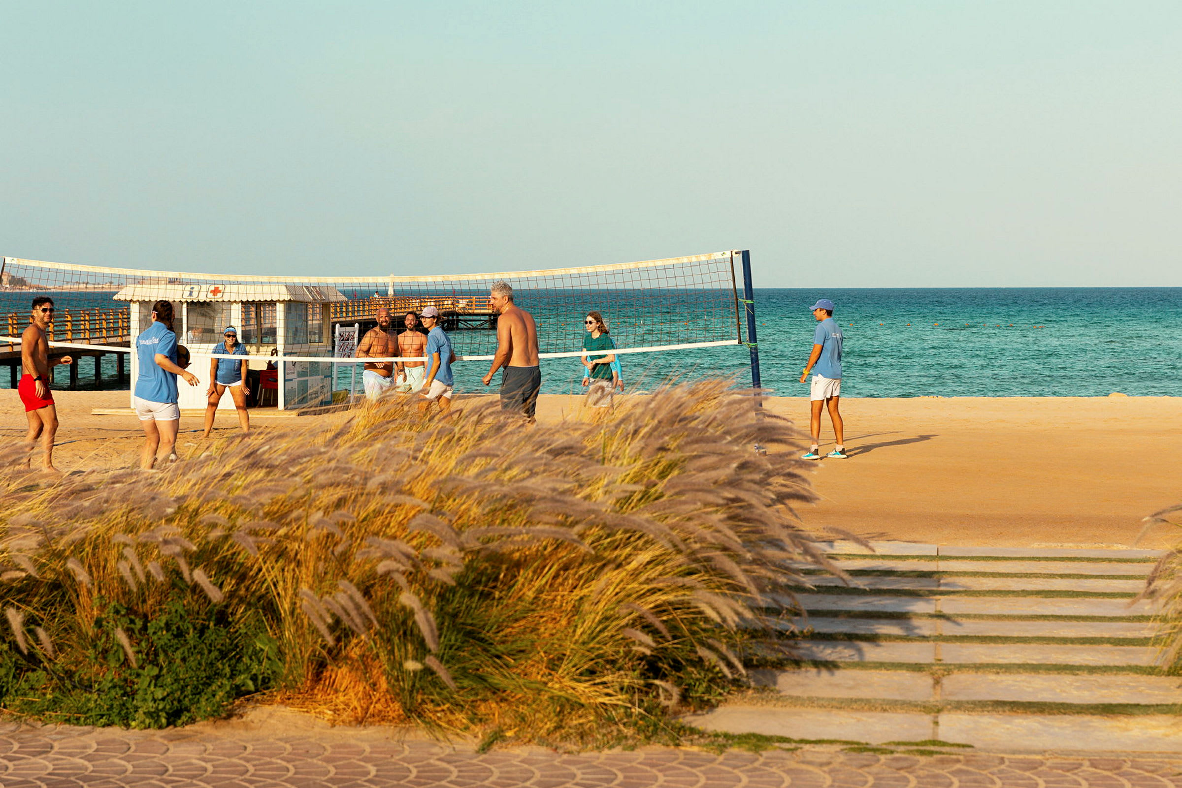 Personer spelar beachvolleyboll vid havet, med gräs i förgrunden. (AI-genererad text)