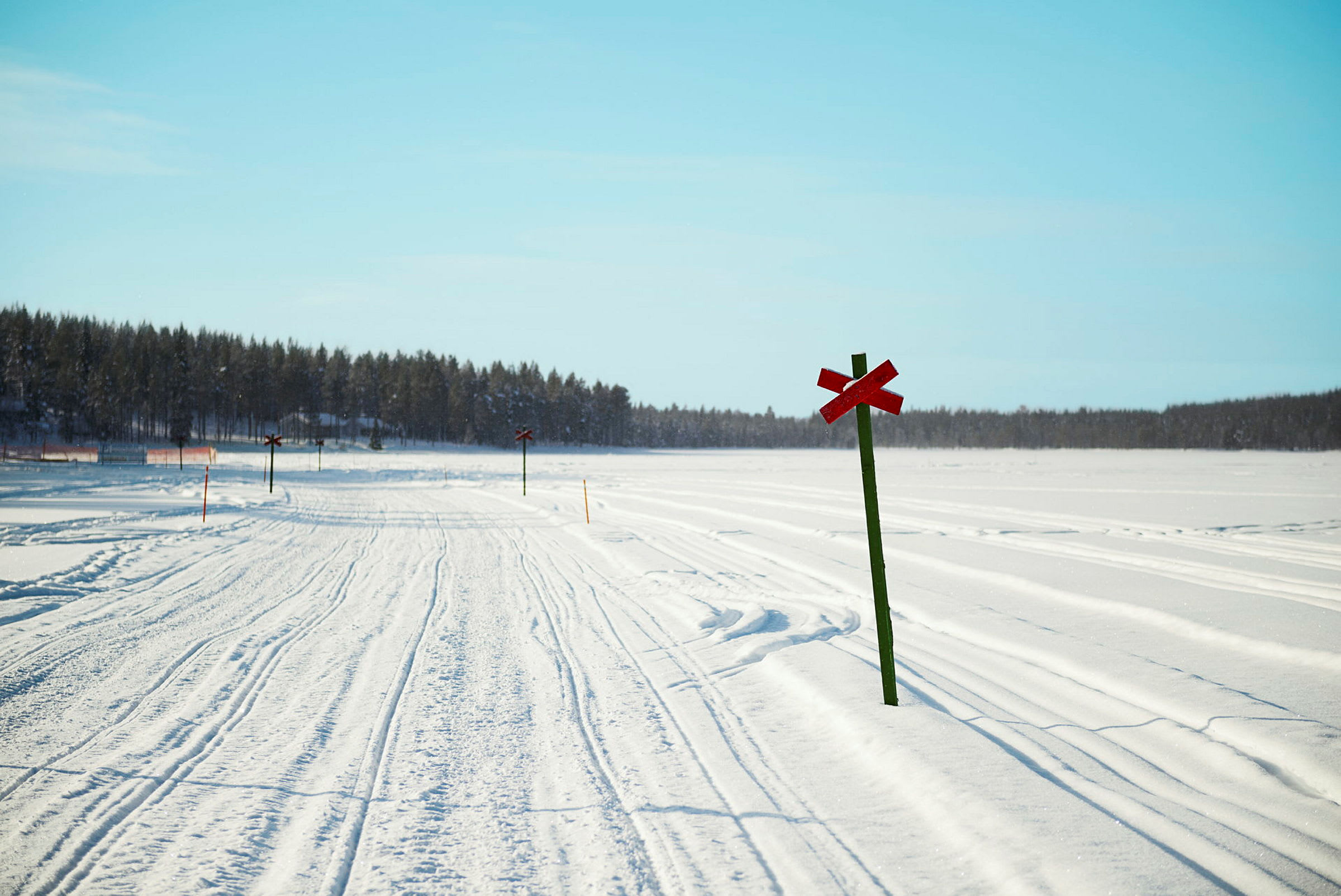 En snöig väg med röda korsmarkerade vägskyltar och skog i bakgrunden under en klar blå himmel. (AI-genererad text)