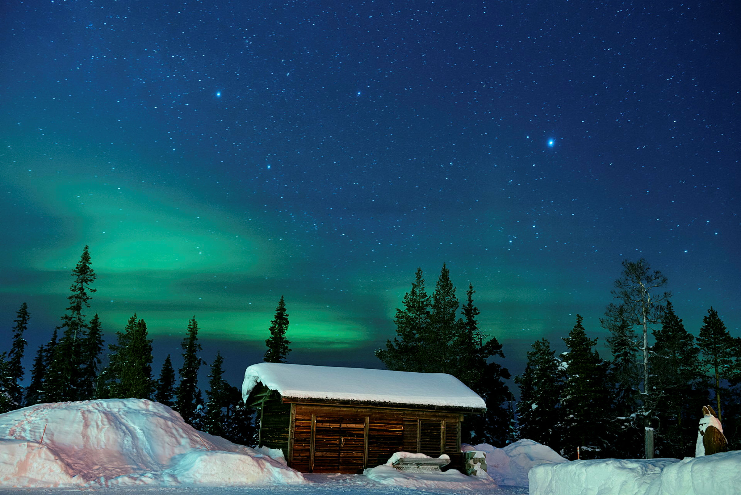 En trästuga med snötäckt tak framför en skog, med stjärnhimmel och norrsken i bakgrunden. (AI-genererad text)