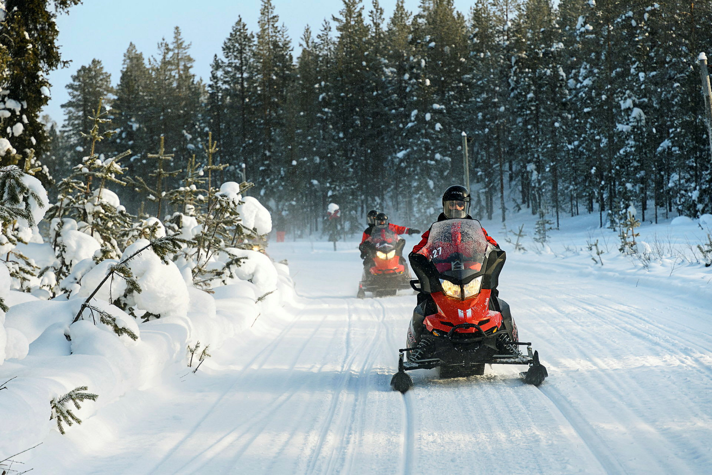 Snöskotrar kör på en snötäckt väg omgiven av skog. (AI-genererad text)