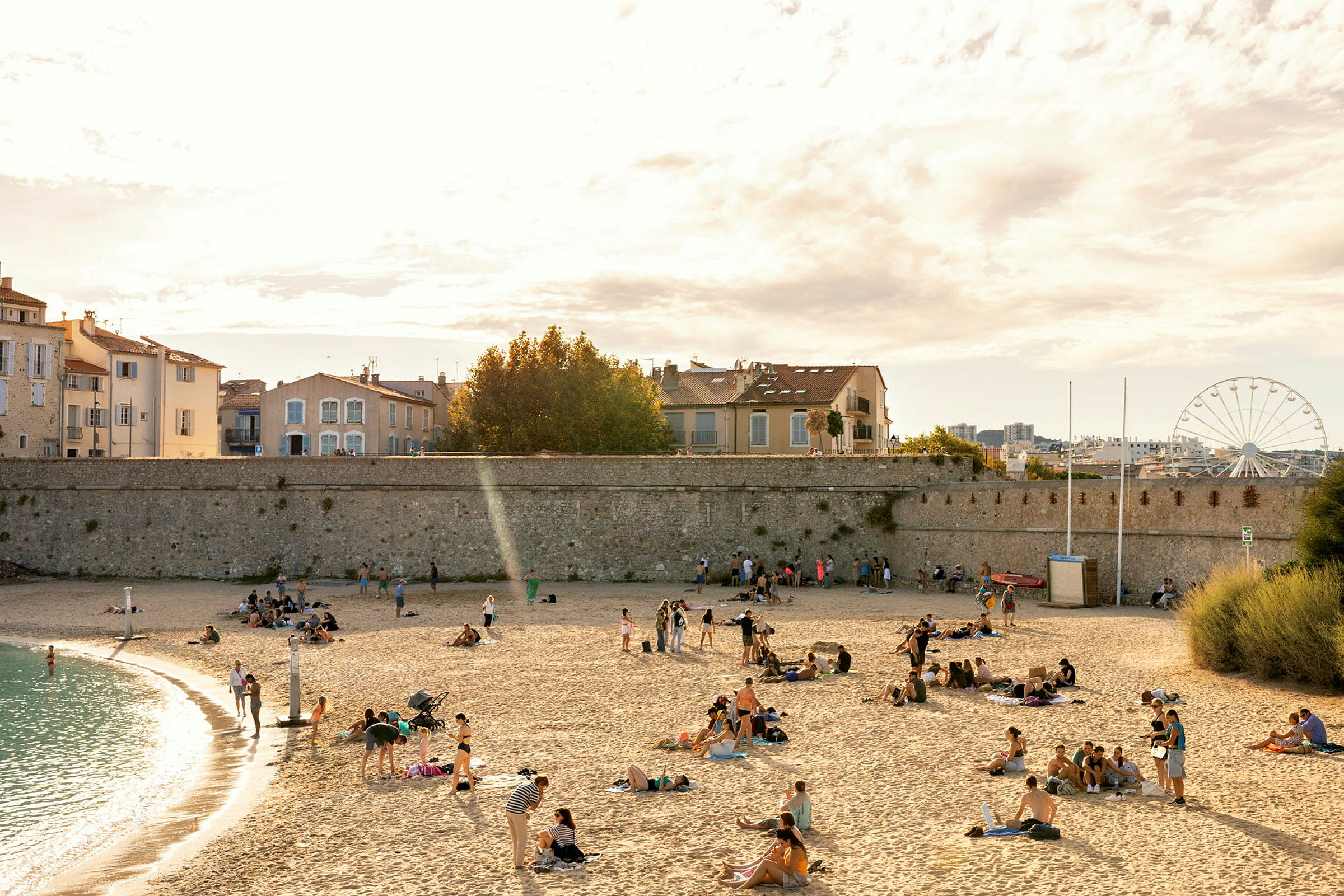 En strand med människor som kopplar av på sanden, medan några individer promenerar vid vattenkanten. Hus och en mur syns i bakgrunden, samt ett pariserhjul. (AI-genererad text)