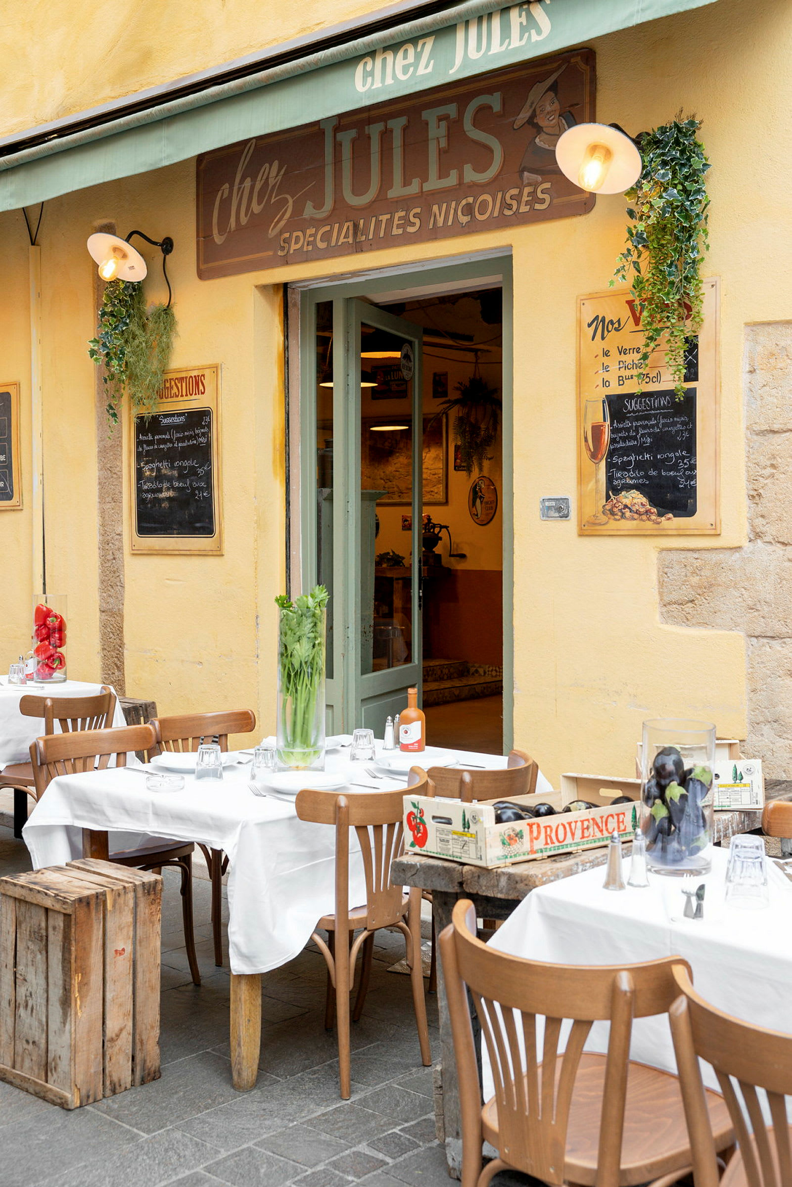 Utomhus sittgrupp med trästolar och bord täckta med vita dukar framför en restaurang med gul fasad och grön skylt. (AI-genererad text)