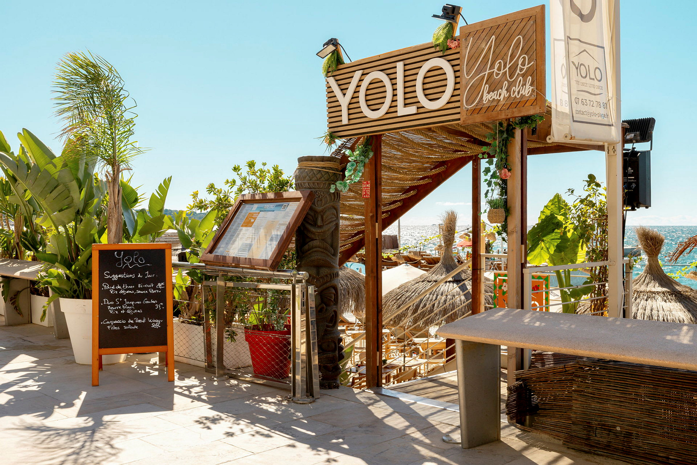 Entrén till en strandklubb med skyltar och grönska. (AI-genererad text)