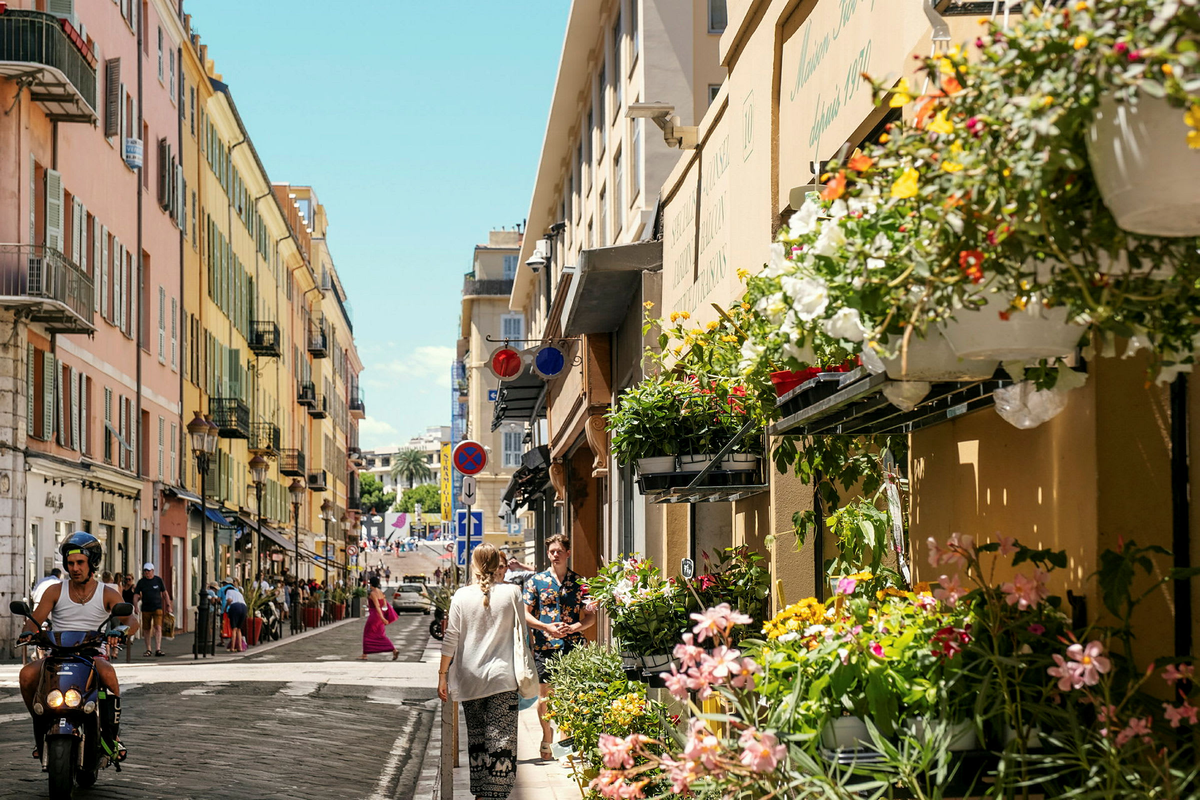 En stadsgata med randiga färgglada byggnader. Blommor är upphängda på fasaden och människor går och åker moped på gatan. (AI-genererad text)