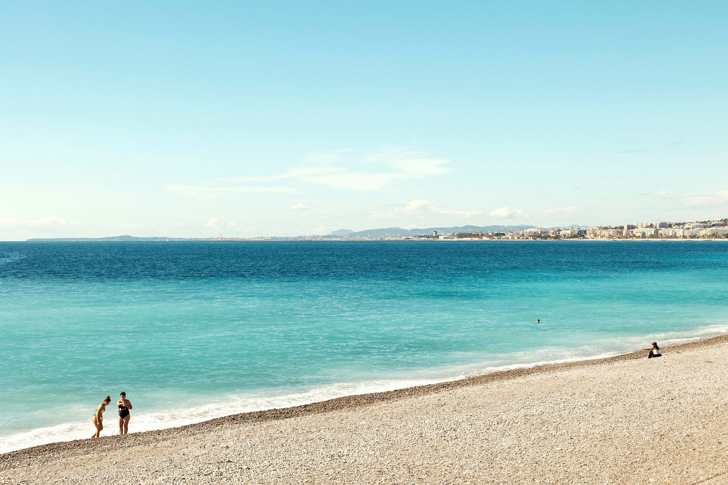 Strand med småsten, hav med blått vatten och en stad i bakgrunden. (AI-genererad text)