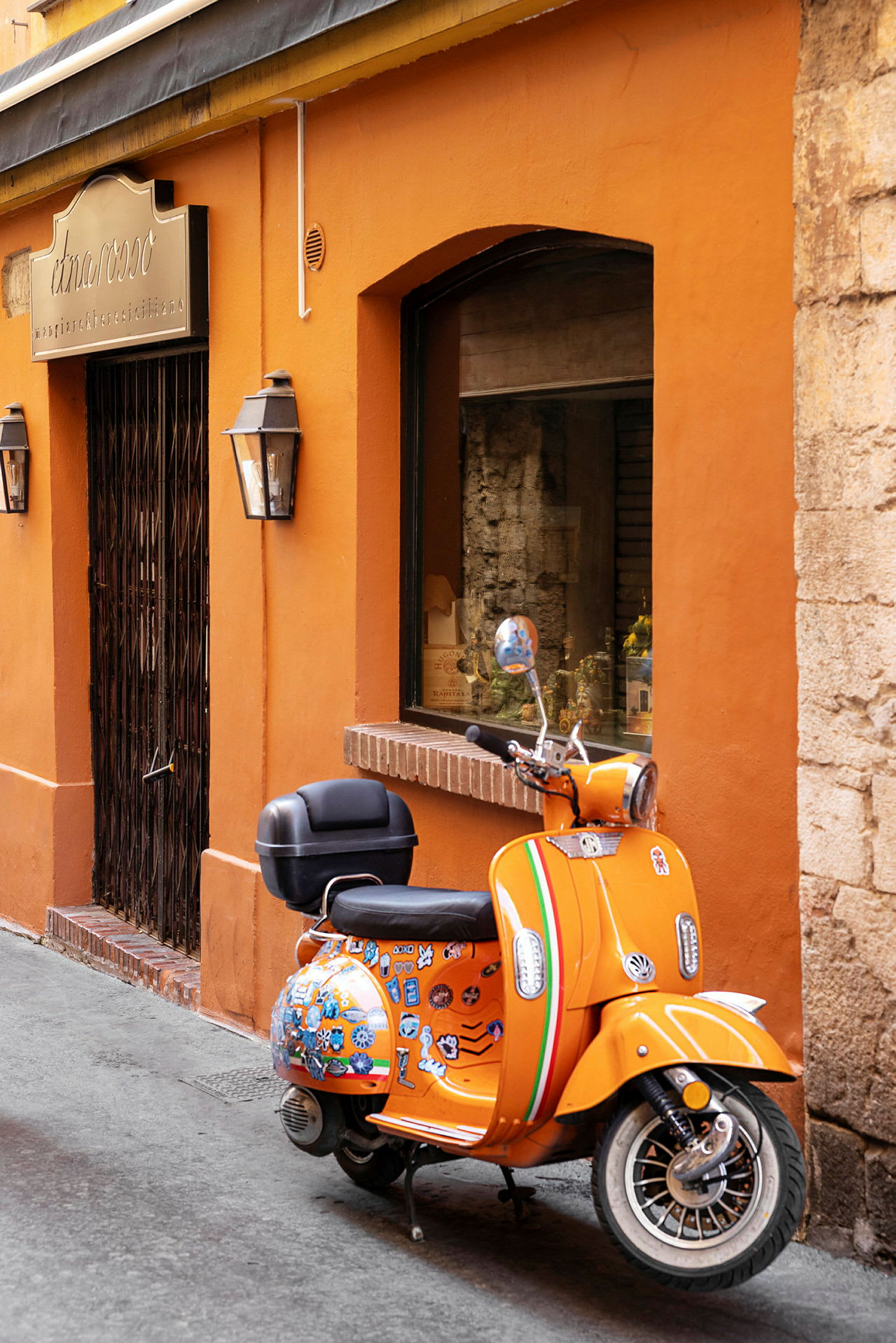 En orange moped med dekorativa klistermärken står parkerad vid en orange byggnad med en smidesjärndörr och ett skyltat fönster. (AI-genererad text)