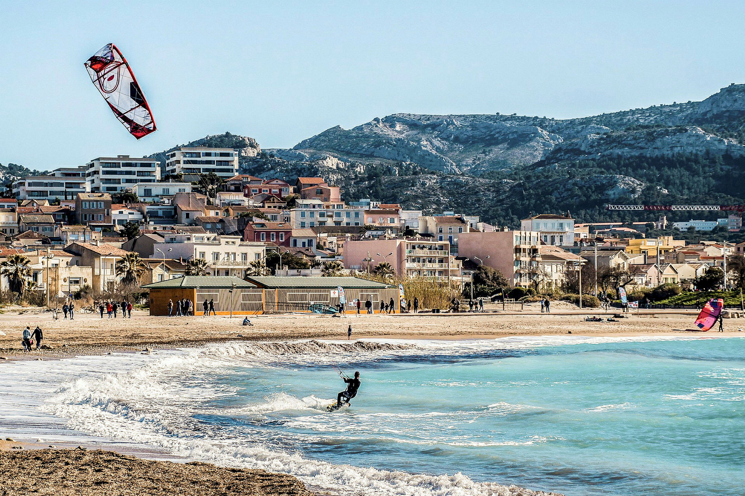 Strand med kitesurfare och vågor, hus synliga i bakgrunden. (AI-genererad text)