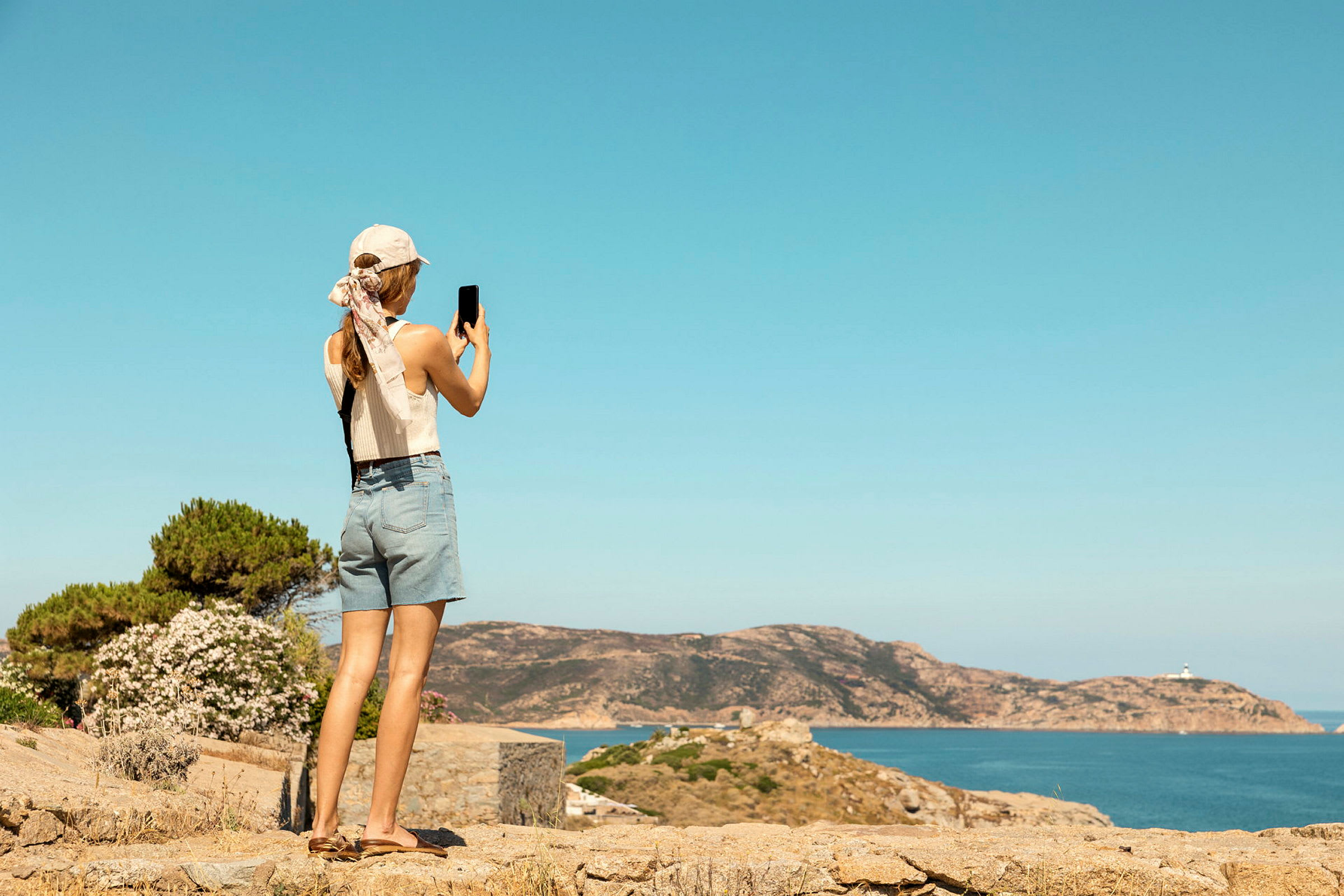 En person med hatt och shorts fotograferar en kustomgivning med hav och klippor. (AI-genererad text)