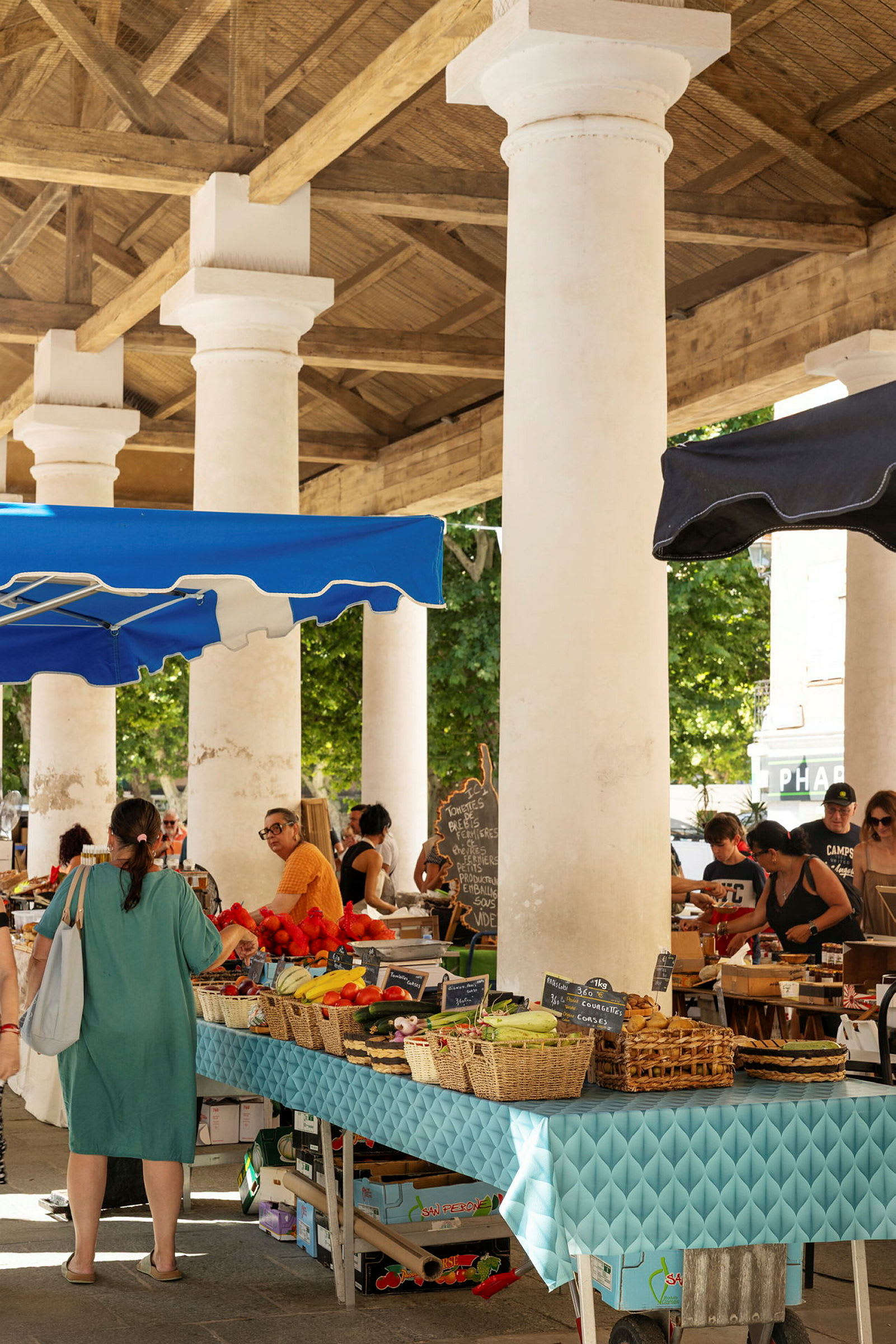 Personer handlar på en utomhusmarknad under en täckt struktur med vita pelare. (AI-genererad text)
